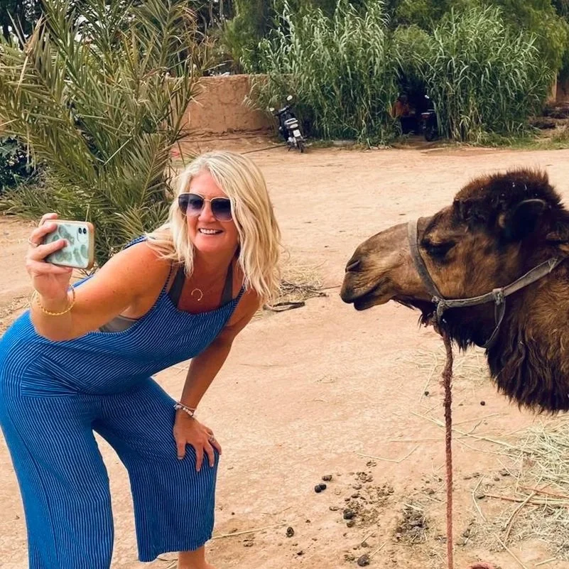A woman with blonde hair, wearing sunglasses and a blue striped outfit, taking a selfie with a camel in a desert setting with greenery in the background.