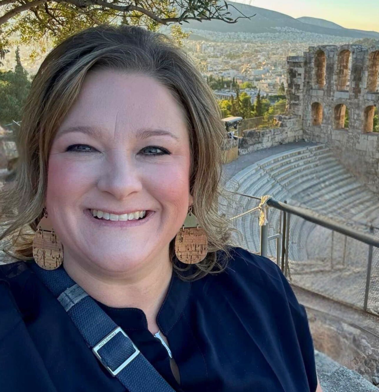A woman smiling in front of an ancient theater with stone seating, with ruins and a cityscape in the background.