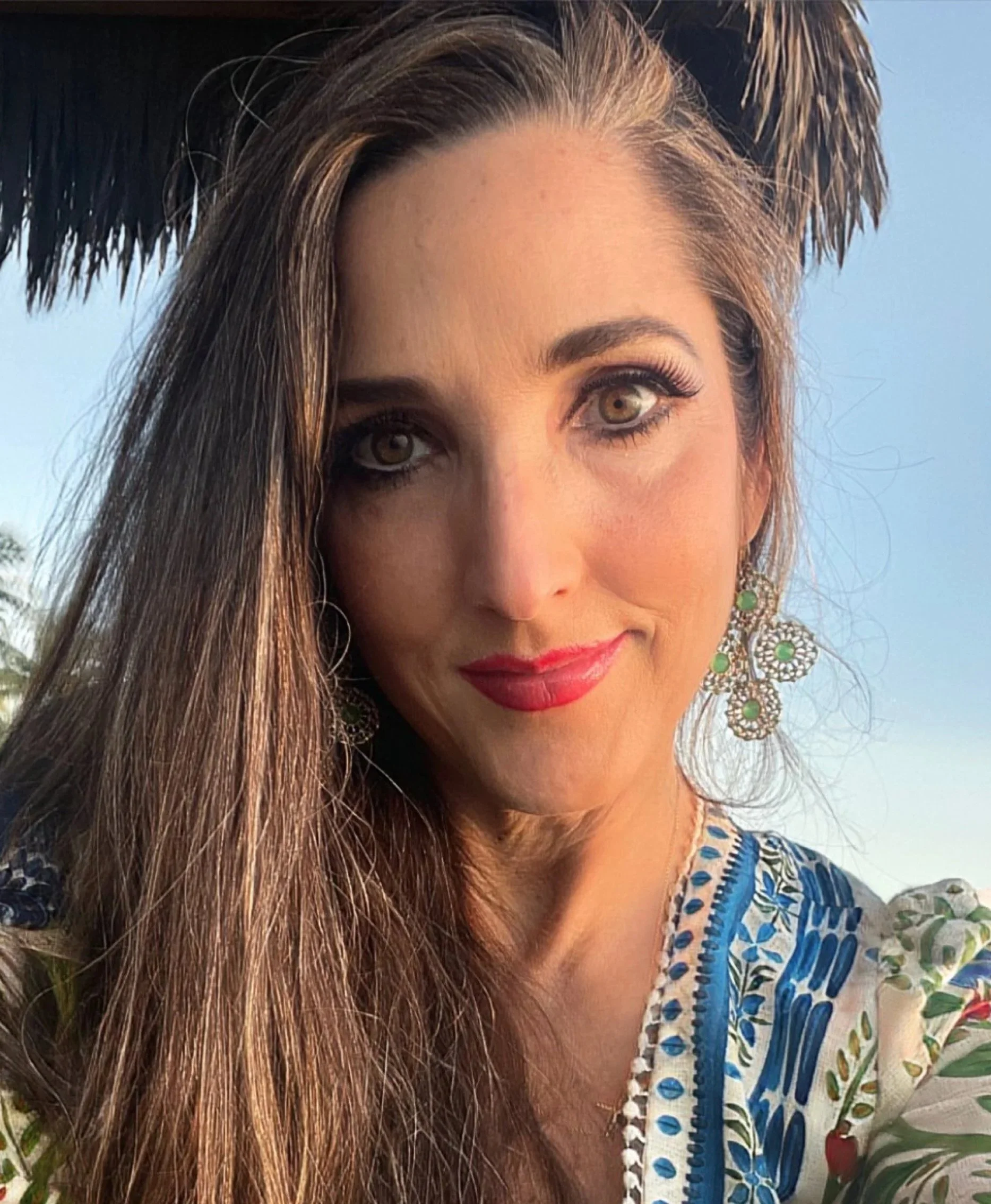 Close-up of a woman with long brown hair, wearing large earrings, red lipstick, and a colorful blue and white patterned dress, outdoors in natural light with a clear sky background.