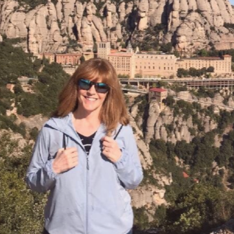 Smiling woman with red hair and sunglasses, wearing a light blue jacket, standing outdoors with rocky cliffs and historic buildings in the background.