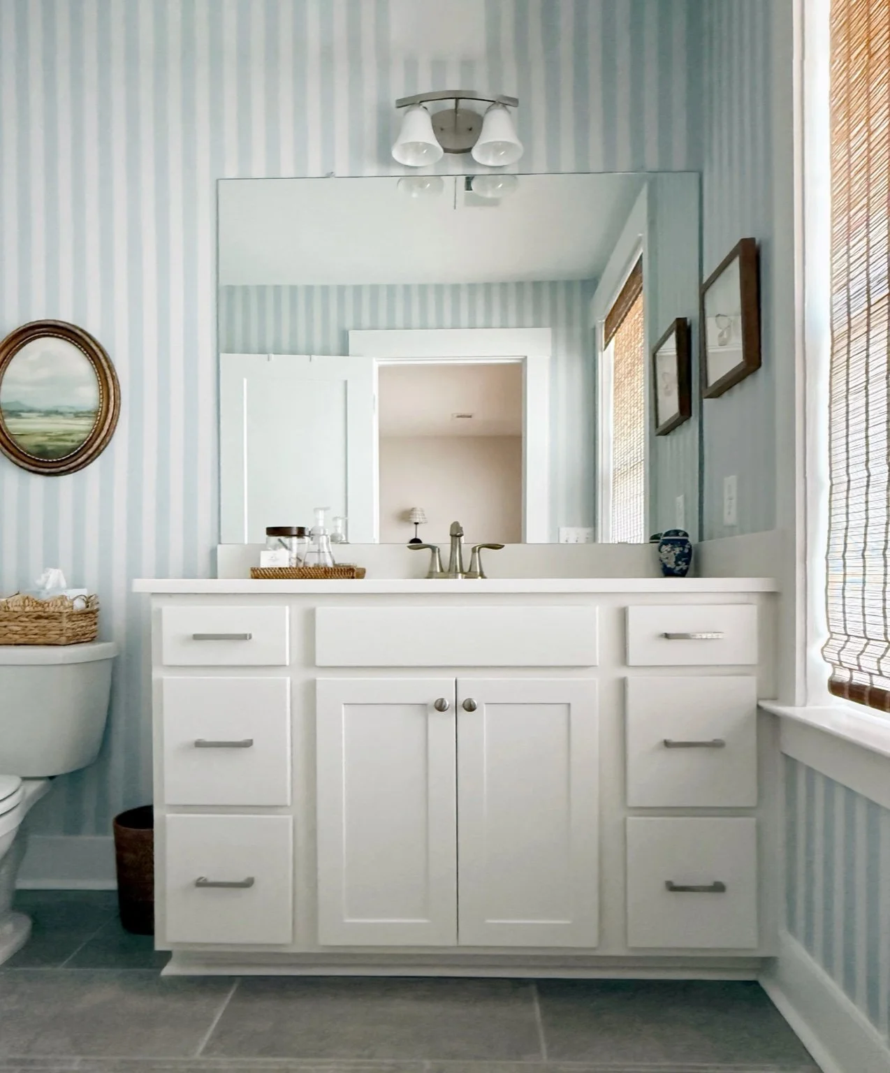 Bathroom with a white vanity, mirror, striped wallpaper, framed pictures, window with bamboo shades, and decorative items on the vanity.