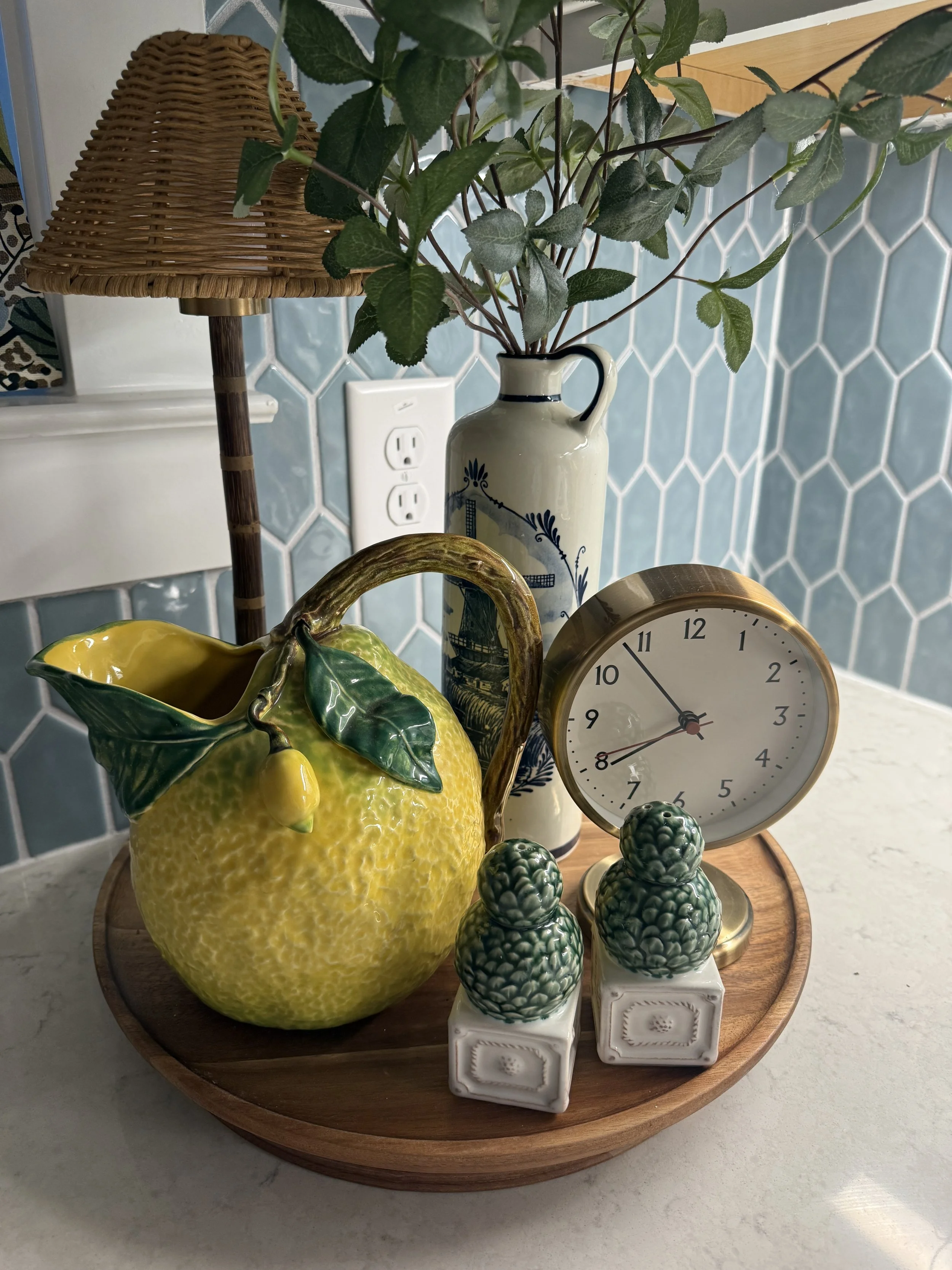 Decorative tray with a ceramic pitcher holding green leaves, a ceramic lemon-shaped container with a clip handle, a vintage-style clock, and three small ceramic pineapple figurines, set on a kitchen counter with a blue tile backsplash and a power out