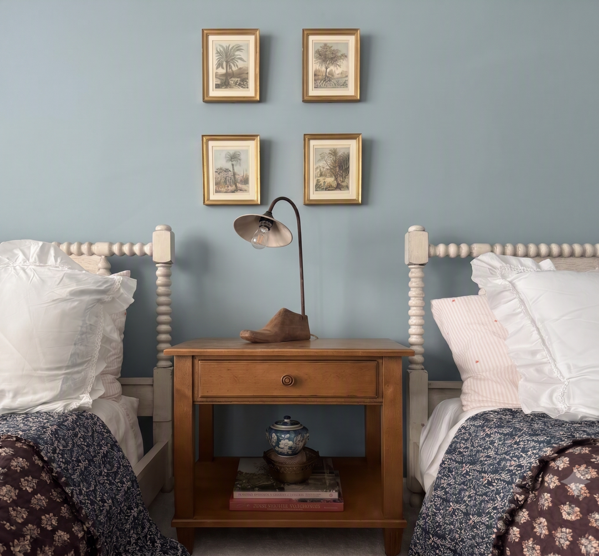 A bedroom with a blue wall and four framed botanical prints arranged in a 2x2 grid. Two beds with white bedding and decorative pillows are on either side of a wooden nightstand. On the nightstand, there is a black and wood table lamp, a wooden sculpt