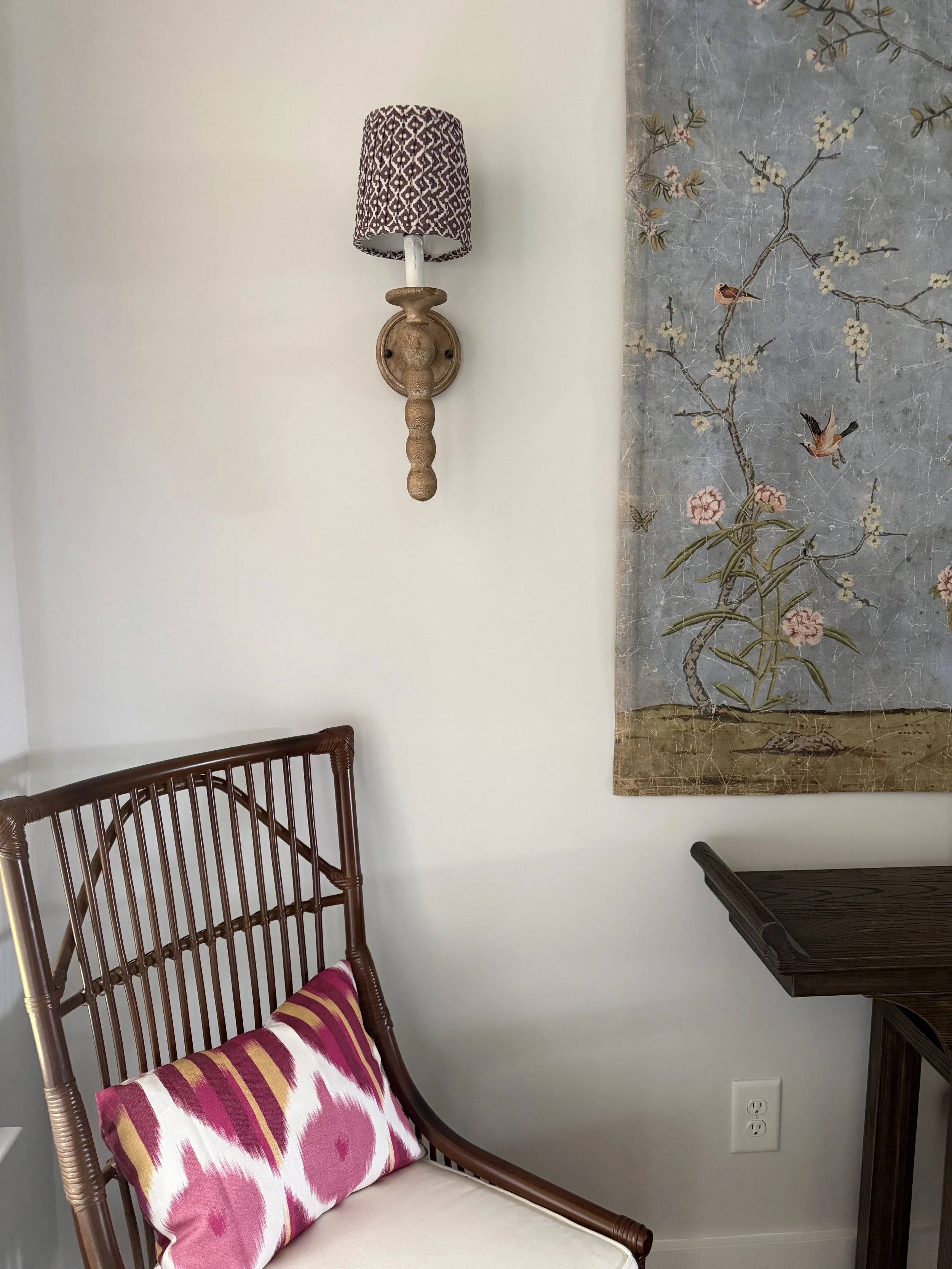 A wooden chair with a colorful pink, purple, and yellow patterned cushion placed on the seat, positioned against a wall. Above the chair, a wall-mounted lamp with a patterned lampshade is attached. To the right, there is a partially visible wooden si