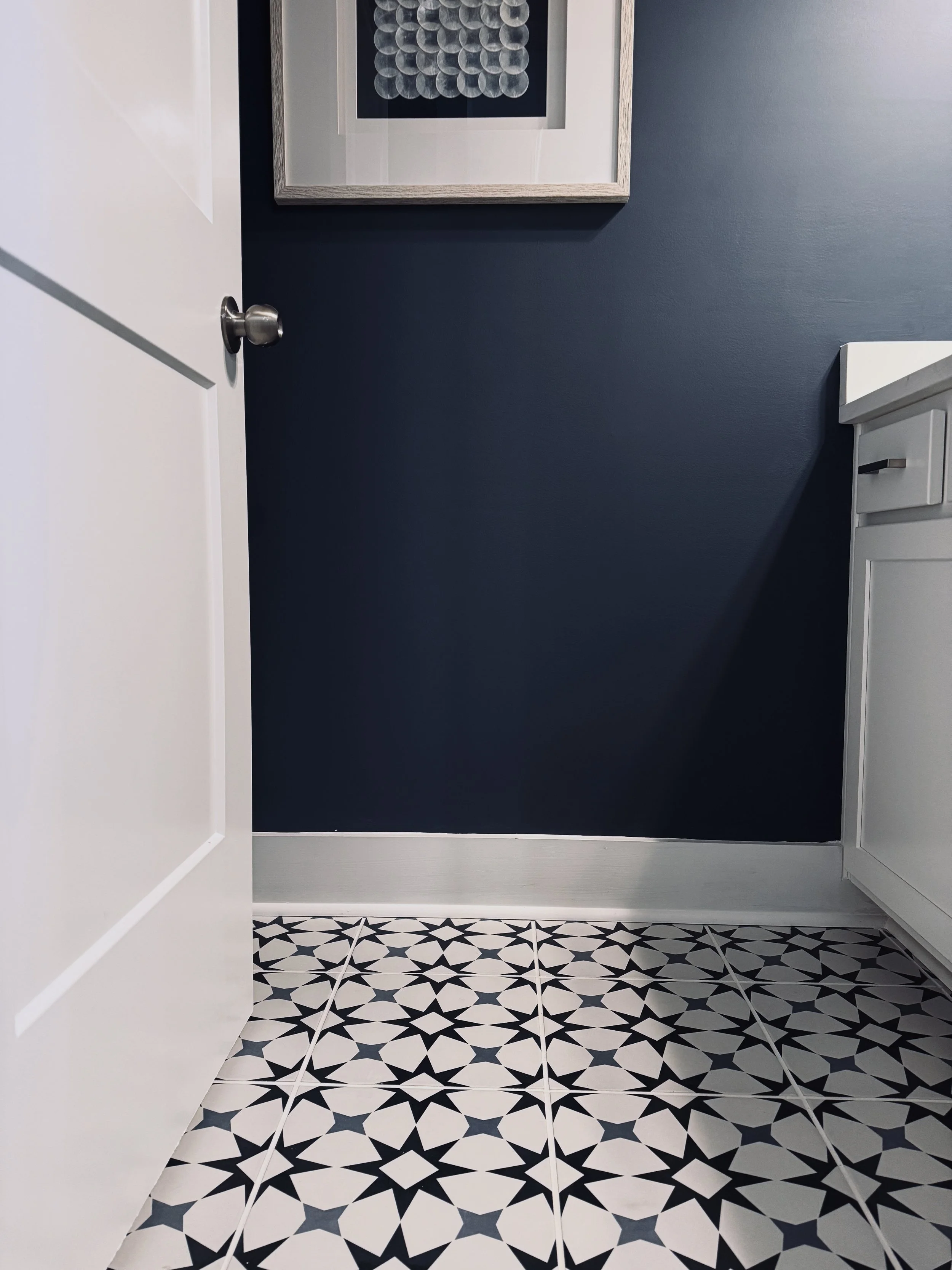 Interior view of a powder room with a navy blue wall, Benjamin Moore Van Deusen Blue, a white door, and a white cabinet. The floor features blue and white patterned tiles, and there is a framed artwork with circular designs hanging on the wall.