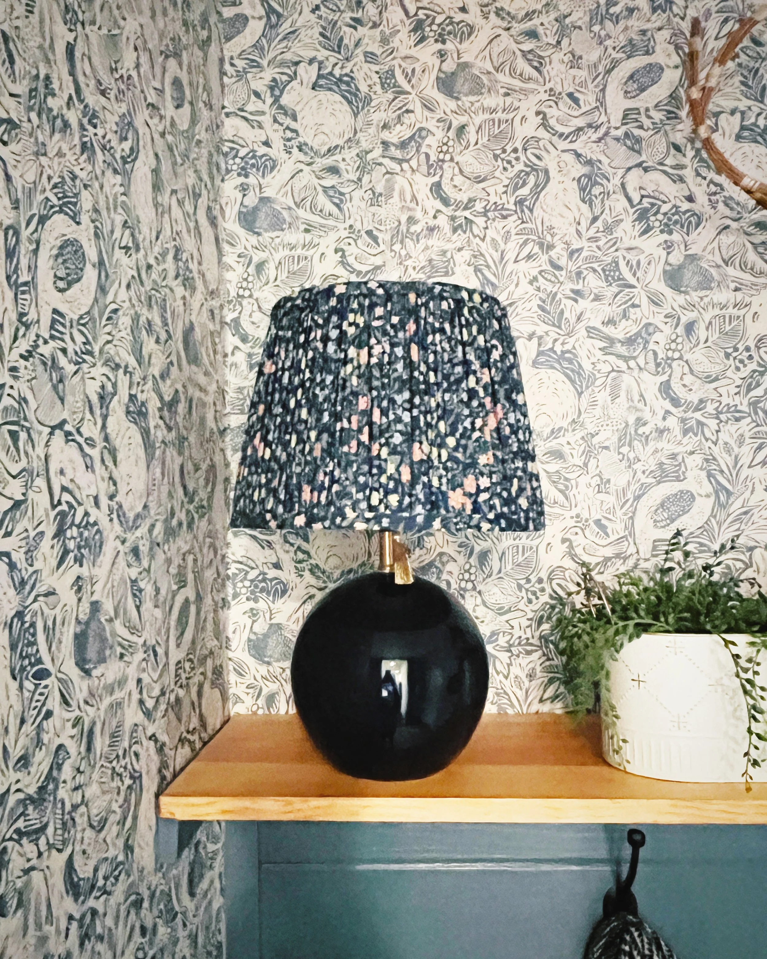 A black round object on a wooden surface, with a gold-colored object inside it, in front of a dark gray door, next to a potted plant with green leaves, and a patterned black and white hanging lamp on the right.