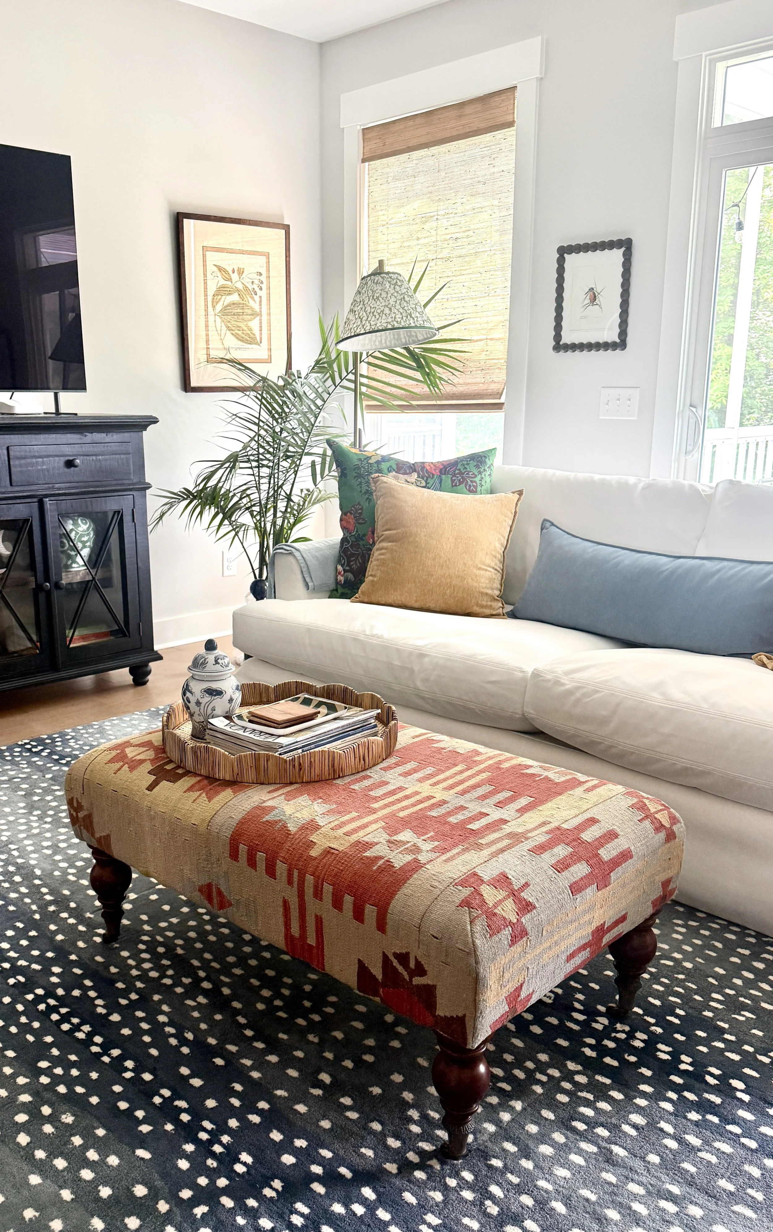 Living room with a white sofa decorated with colorful pillows, a patterned ottoman with a tray and magazines, a dark wooden cabinet, framed artwork on the walls, a large window with bamboo blinds, a tall potted plant, and a patterned area rug.