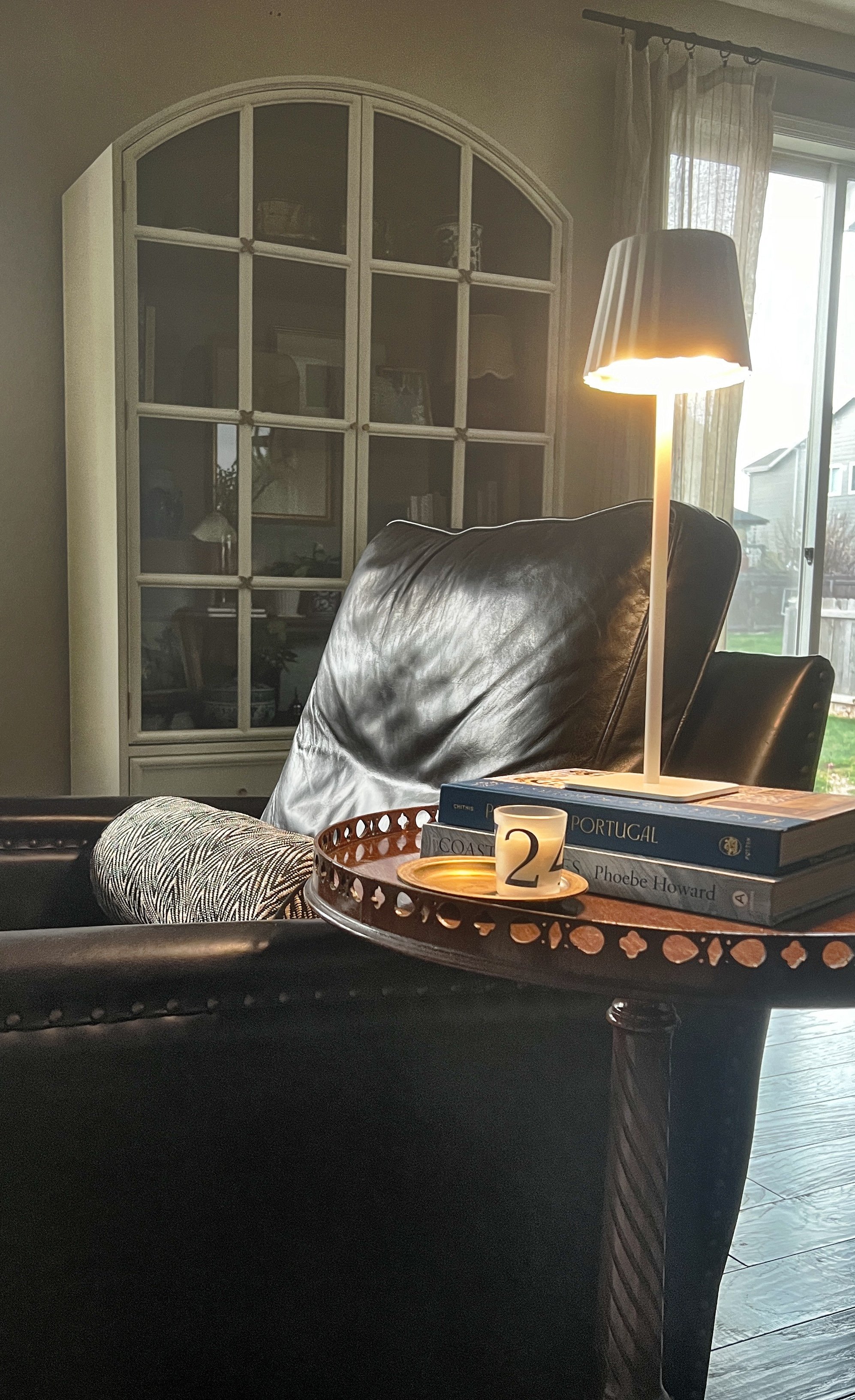 A living room with a black leather armchair, a lamp, a tray with a candle, a stack of books, and a window with curtains.