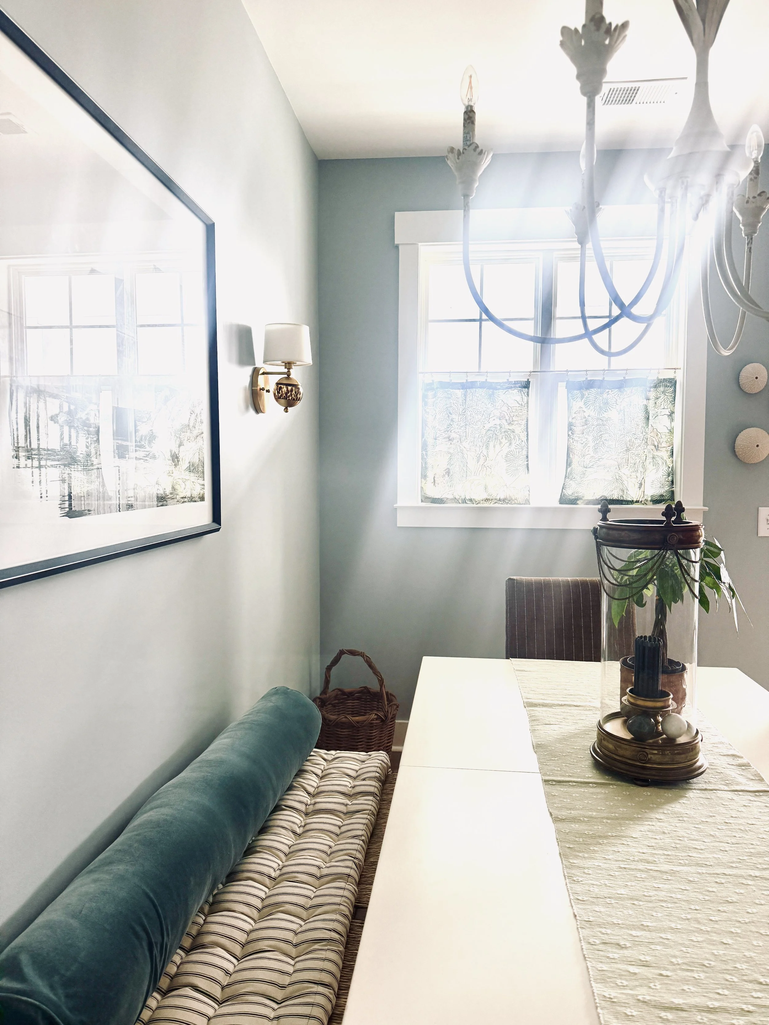 Corner of a dining room with a window, curtain, decorative wall art, a chandelier, a wall-mounted lamp, a dining table with a table runner, a glass vase with plants, and chairs.