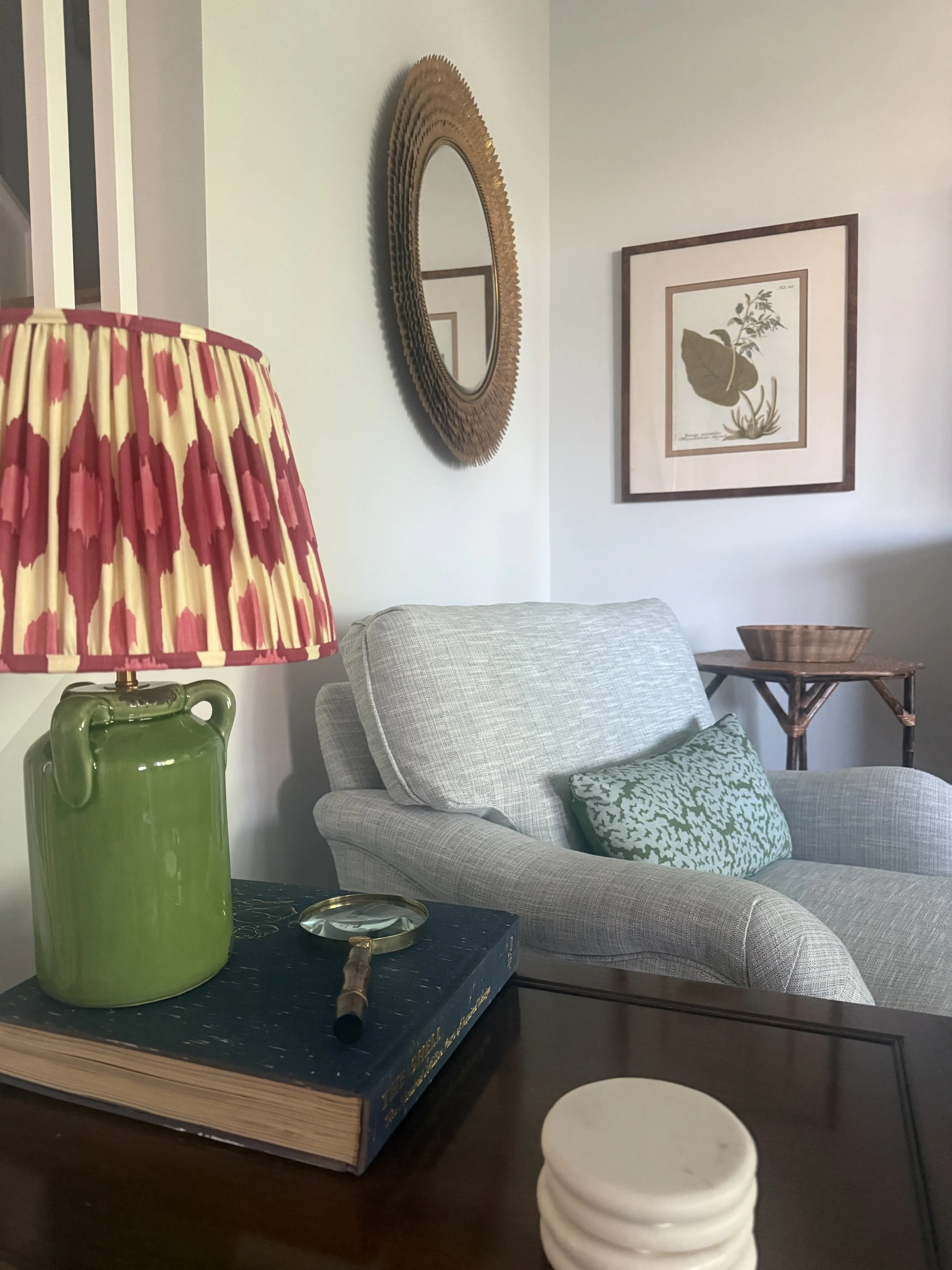 Close-up of a black table with a large green ceramic vase, a stack of white coasters, a box with a magnifying glass on top, a backpack, a light gray armchair with a green patterned pillow, a wooden side table with a woven basket, framed botanical pri