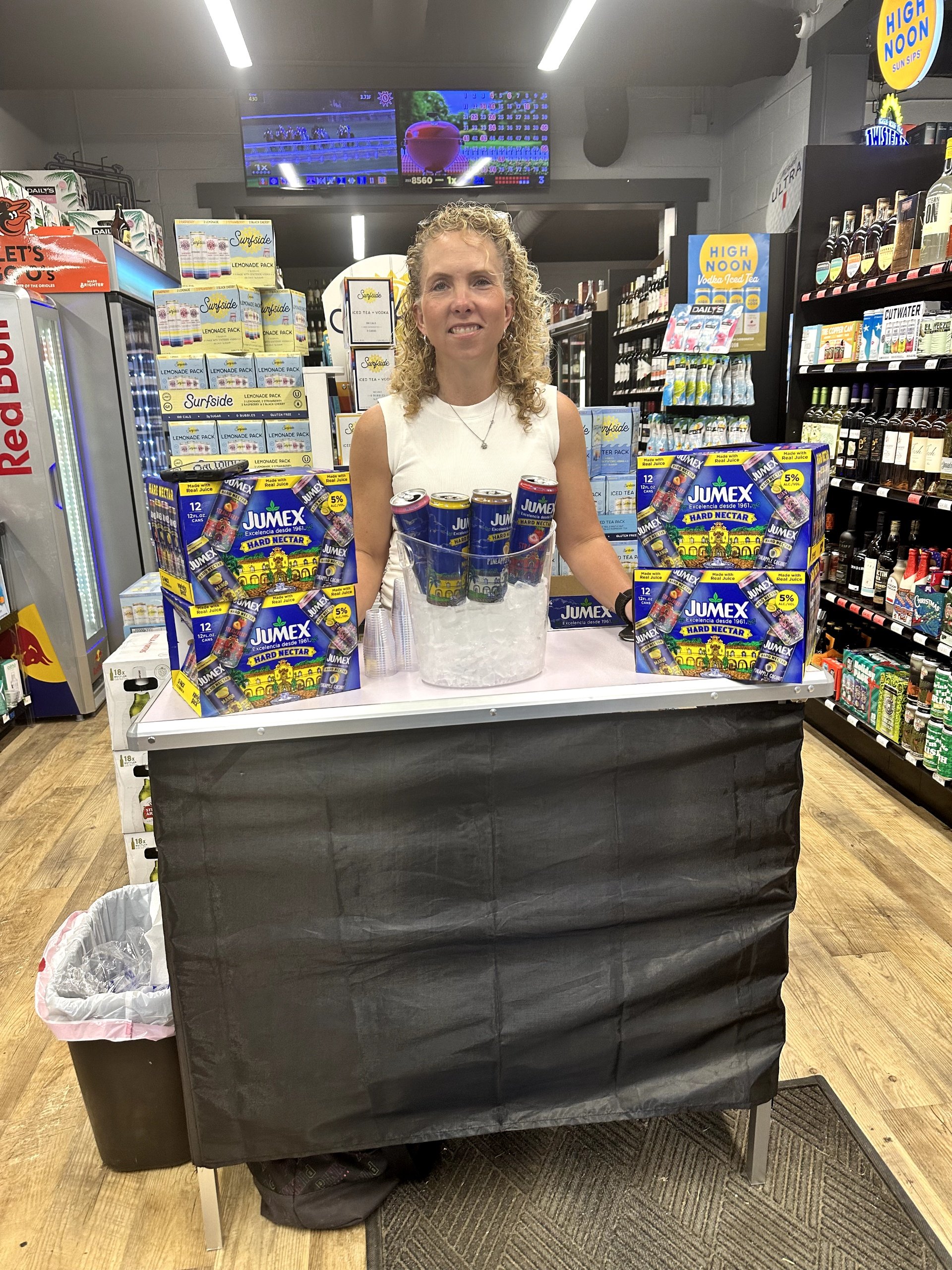 A woman standing behind a small lemonade stand inside a grocery store, with cans of Jumex juice and a bucket of ice in front of her, with a grocery aisle and a digital screen above in the background.