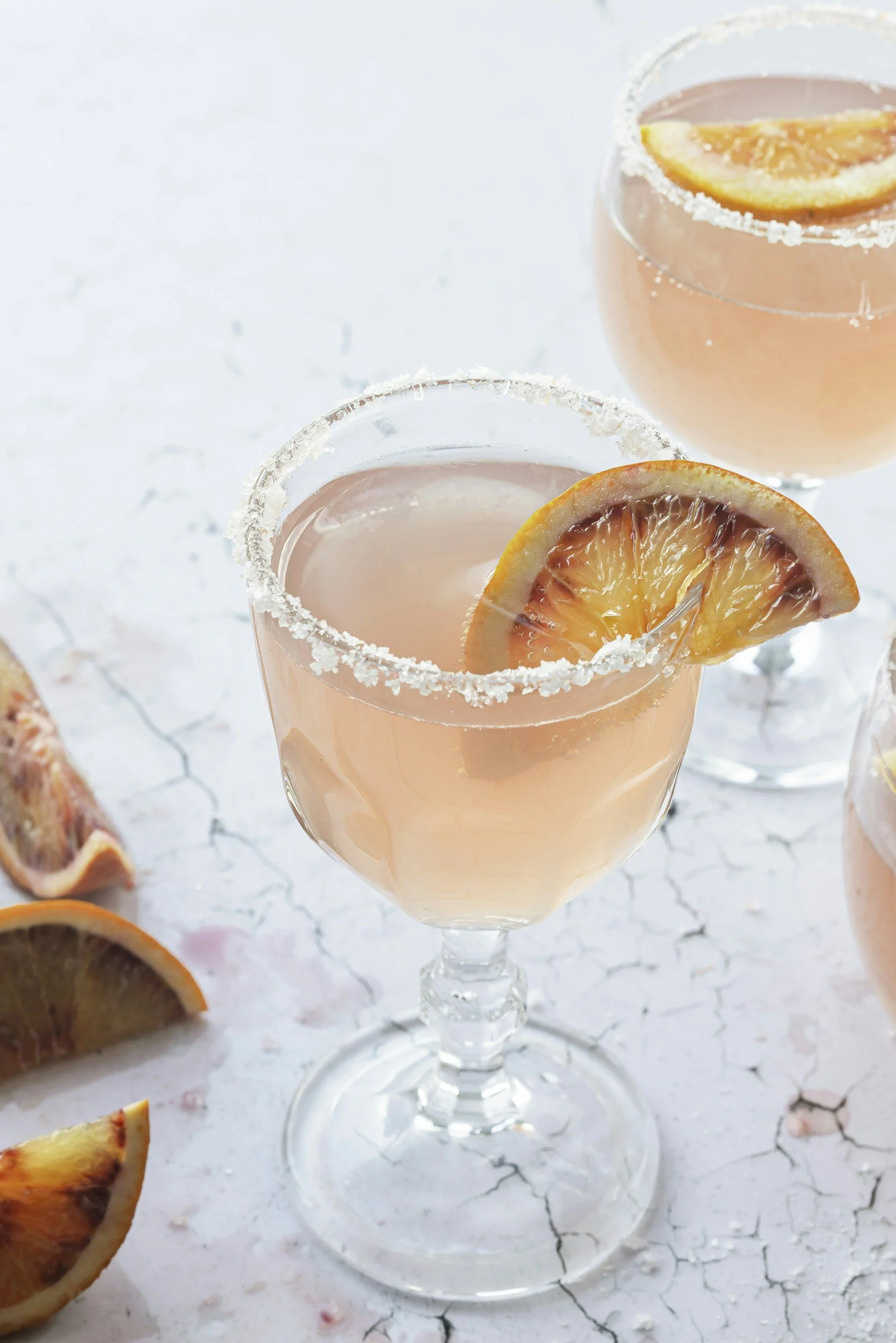 A glass of pink cocktail garnished with a blood orange slice and rimmed with salt, with additional blood orange slices surrounding it.