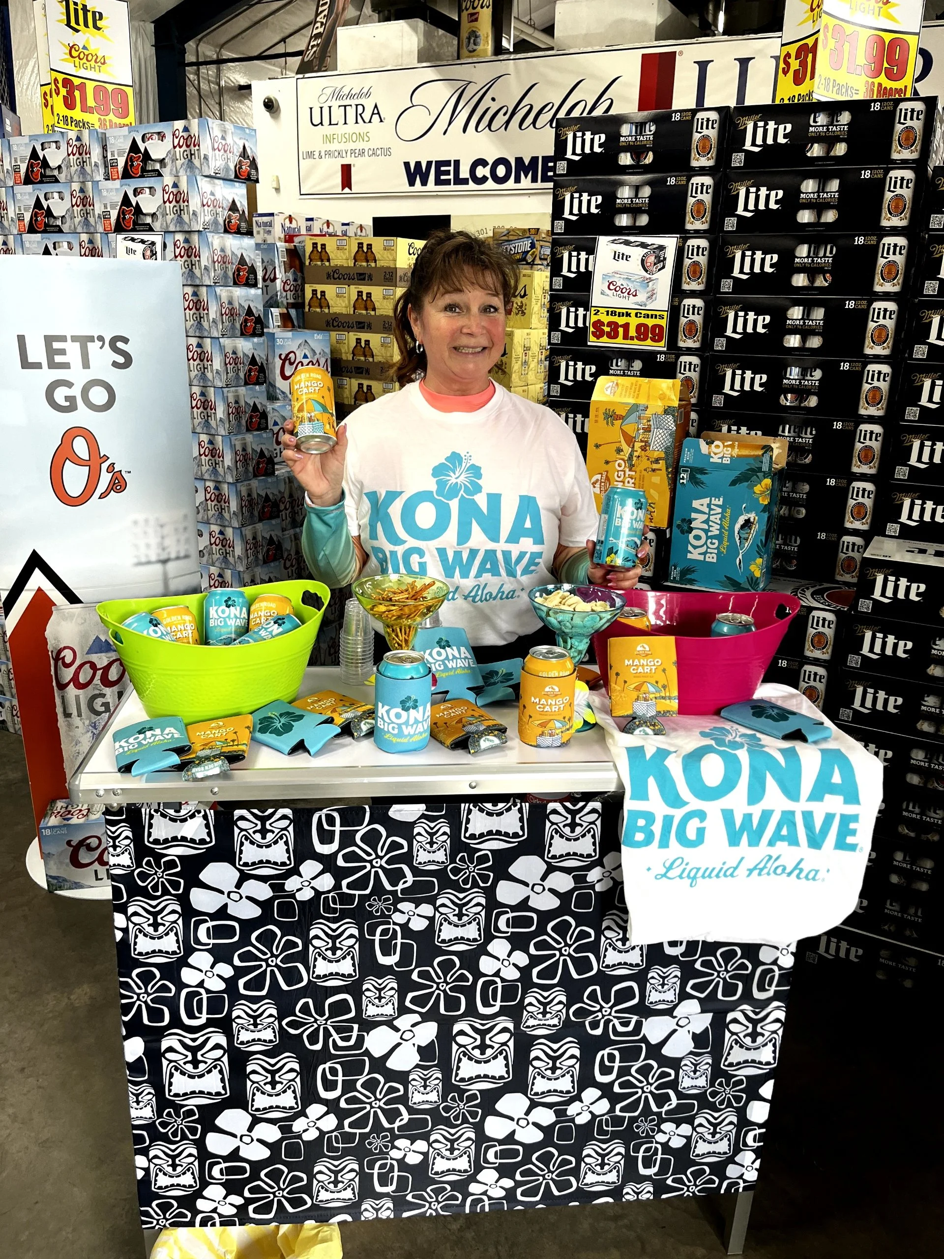 Woman smiling at a booth promoting Kona Big Wave beer with cans and merchandise, with a busy store background of beer cases and signage.