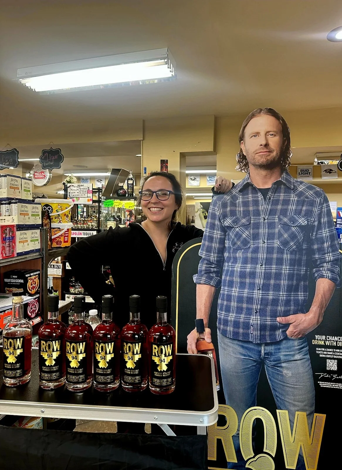 A woman with glasses and dark hair, smiling, stands next to a cardboard cutout of a man in a plaid shirt holding a bottle. In the foreground, bottles of 'ROW 94' whiskey are displayed on a table.