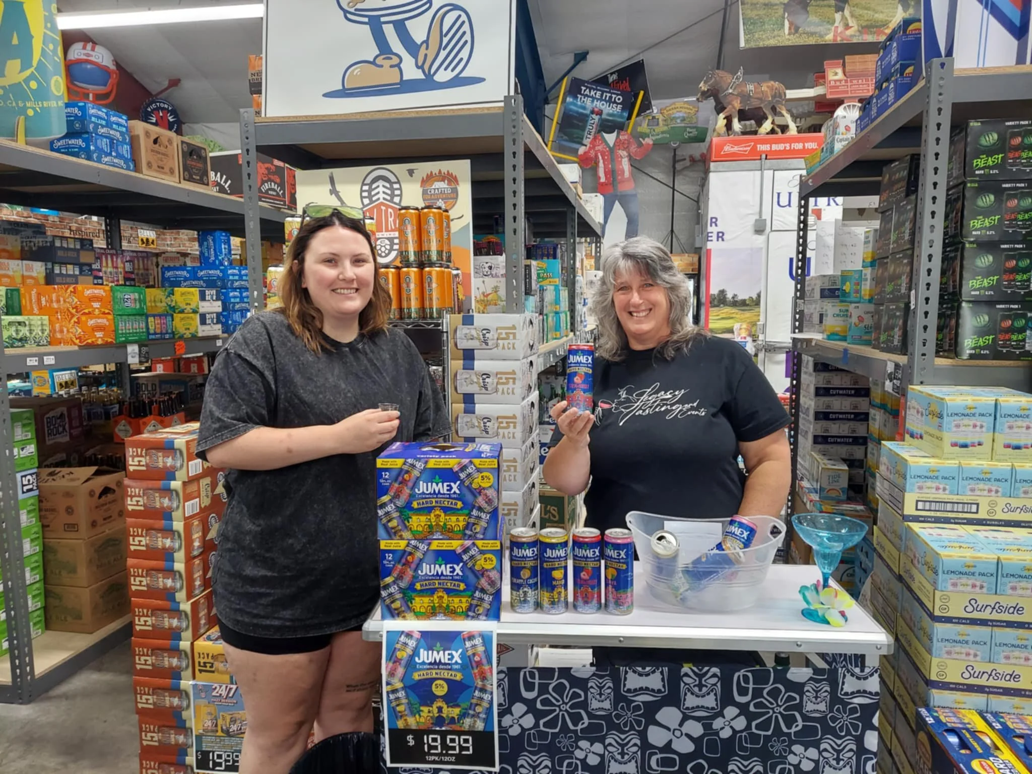 Two women standing at a product booth in a store aisle, holding cans of Jumex nectar and smiling. Shelves stocked with various products surround them, and promotional signs are visible.