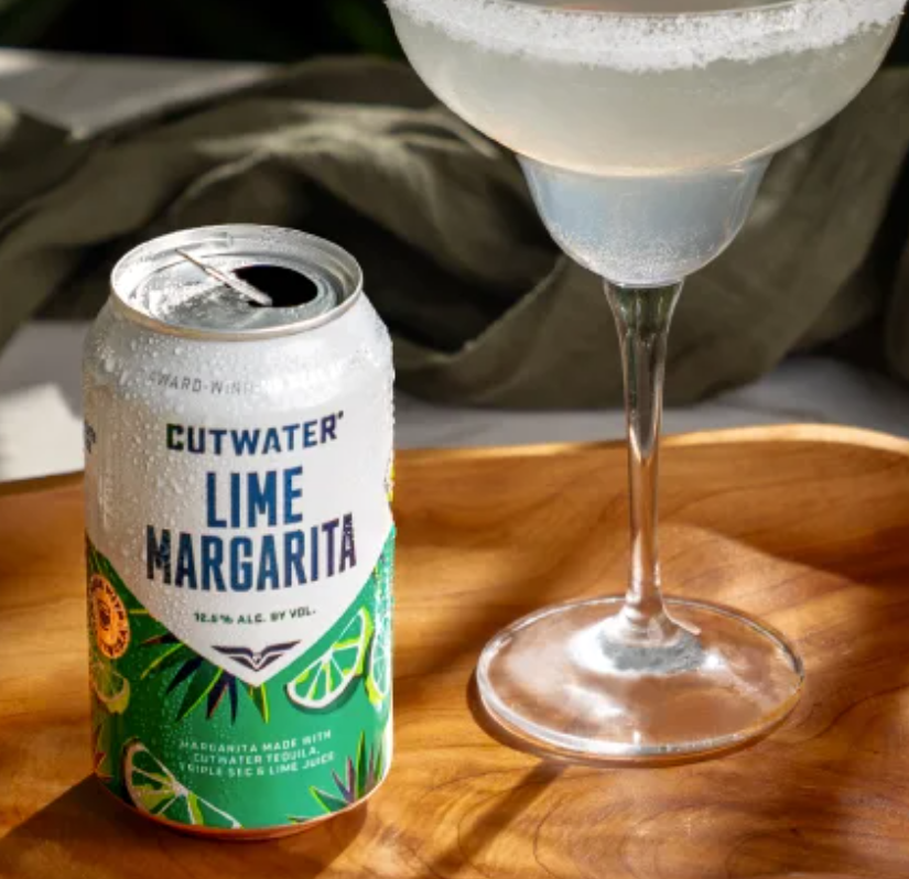 A can of Cutwater Lime Margarita with an open top, next to a glass of margarita in a stemmed glass. The can has a green, yellow, and white label with lime illustrations. The drink appears frosty, on a wooden tray.