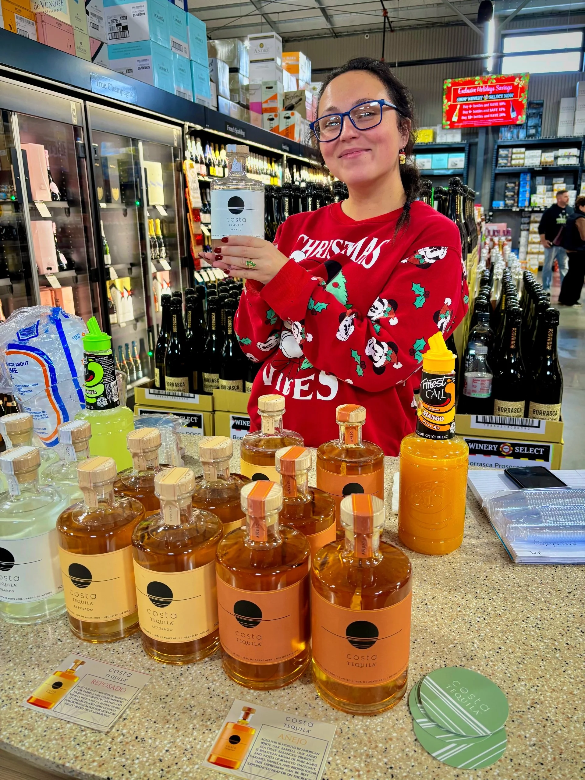 A woman in a red Christmas sweater with festive prints stands at a store display of Costa Tequila bottles, holding a large bottle of tequila. The display features multiple bottles on a countertop along with promotional cards and a yellow liqueur. In 