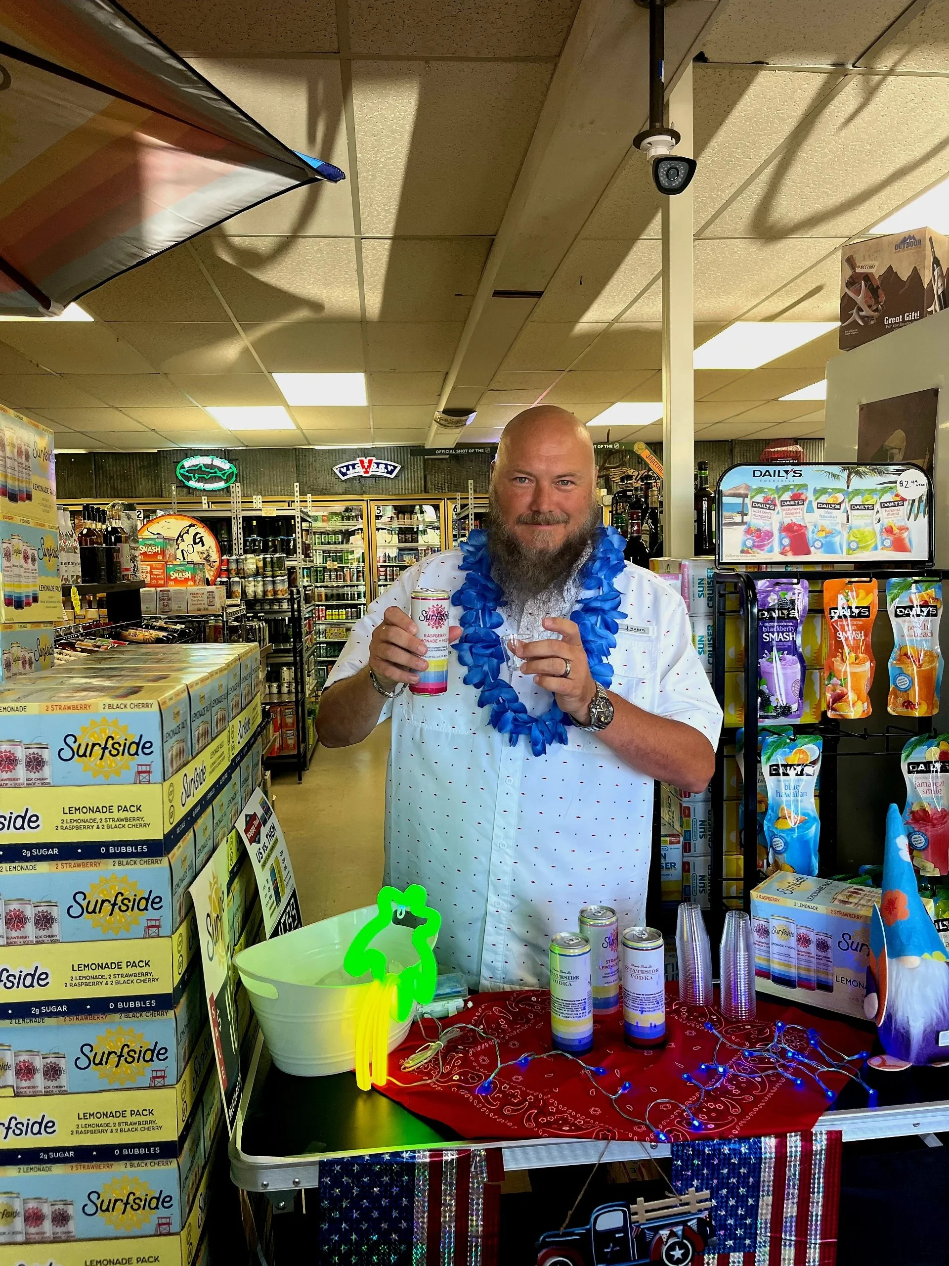 Man with a beard wearing a white shirt with red dots, blue lei, and a watch, standing behind a table with beverage cans, holding a can and a shot glass. The table has party decorations, a neon sign shaped like a bear, and is set in a convenience stor