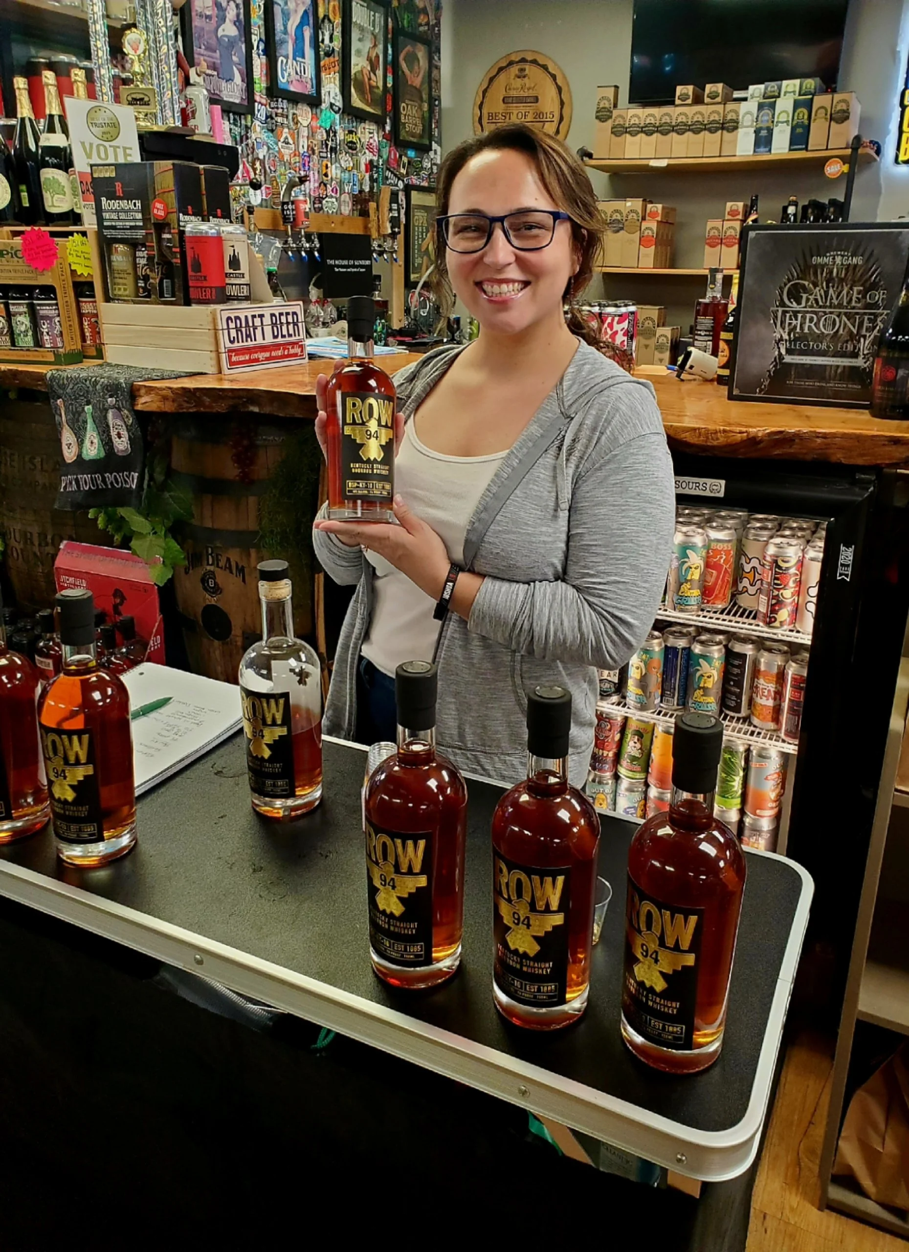 A woman smiling and wearing glasses, holding a bottle of ROW 94 whiskey, standing behind a table with several bottles of the same whiskey on display, in a store with shelves of drinks, craft beers, and decorative items.