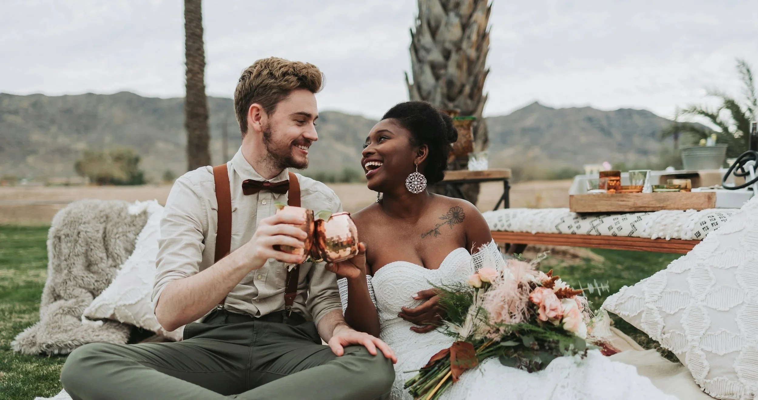 A couple at a wedding or celebration, sitting outdoors on grass with mountains in the background, raising copper mugs in a toast and smiling at each other. The woman wears a strapless white dress and large earrings, with a bouquet of flowers beside her.