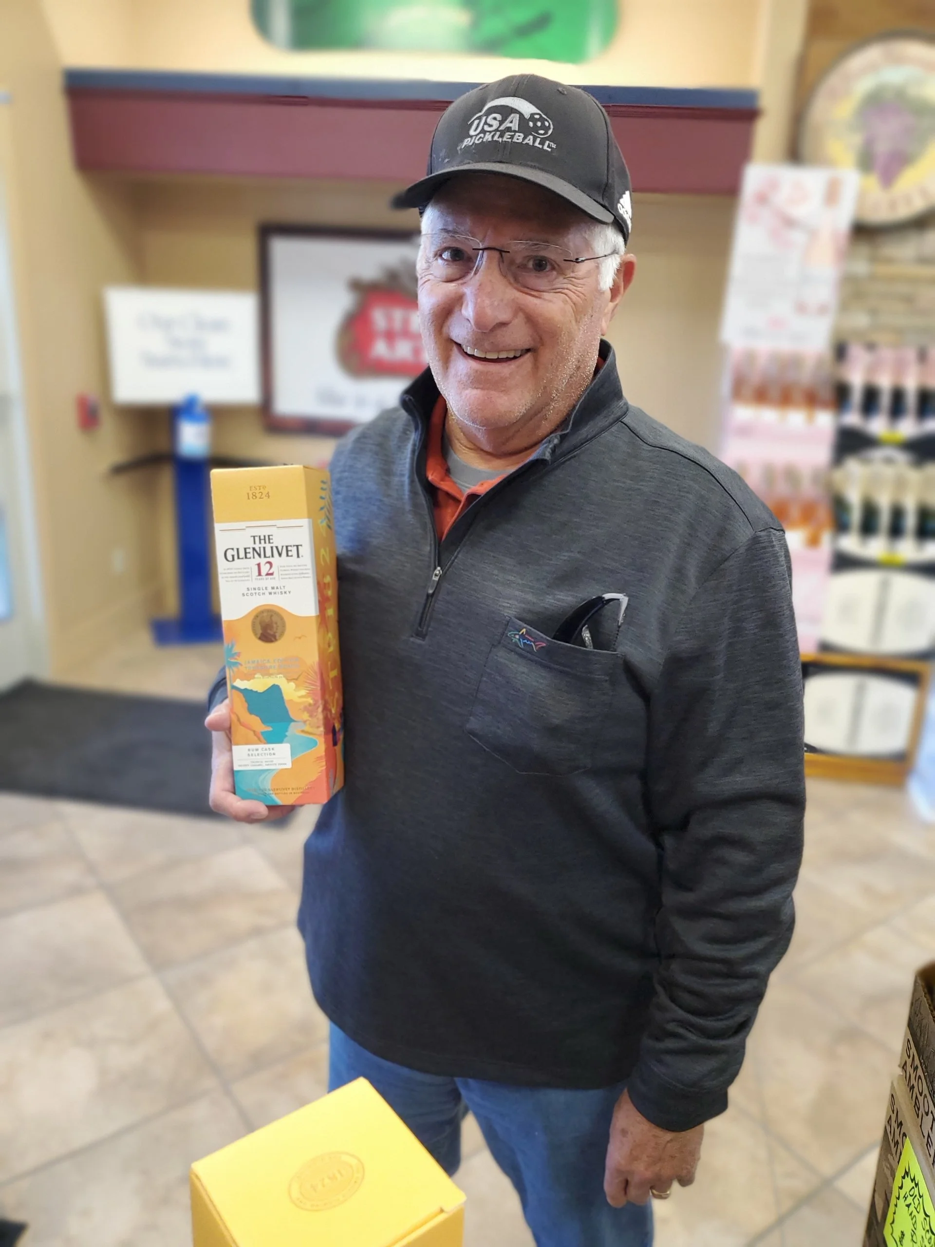 A smiling man wearing glasses, a black cap with 'USA PICKLEBALL' on it, and a gray zip-up jacket, holding a box of The Glenlivet 12 Scotch whisky inside a store.