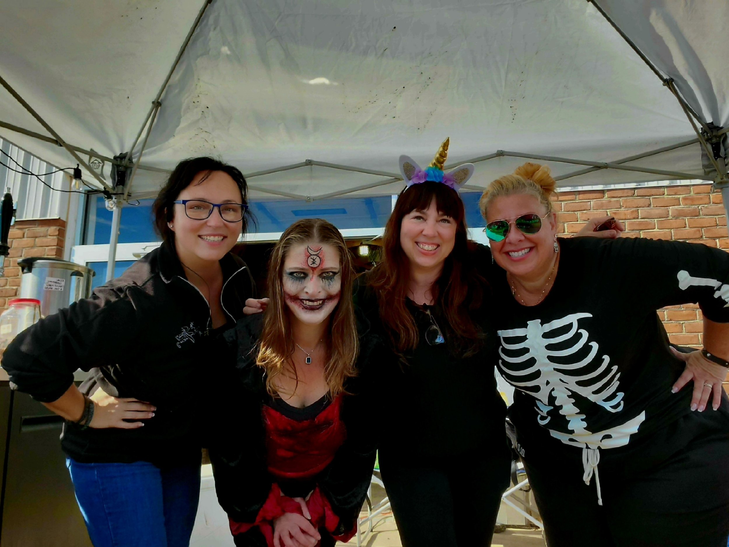 Four women smiling and posing together under a canopy, with one woman dressed in a unicorn headband and costume, and another in a skeleton shirt, possibly at a Halloween or costume event.