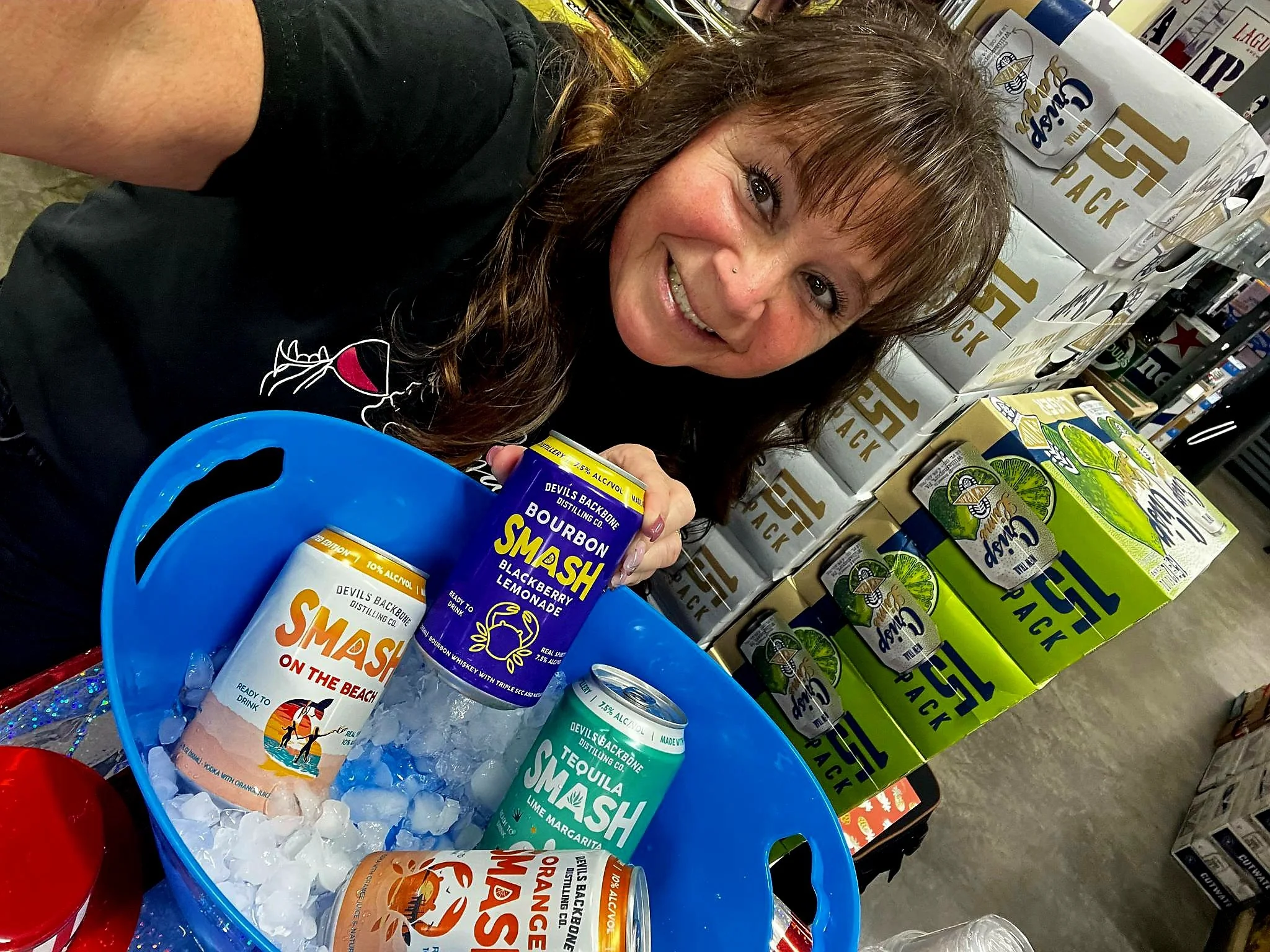 A woman smiling at a store display, holding a can of Smash blackberry lemonade. She is surrounded by boxes of Smash beverages and a blue ice bucket containing cans of Smash drinks, including tequila lime margarita, on the beach, and orange margarita.