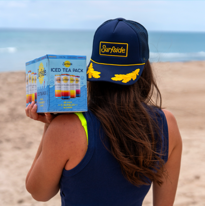 A woman at the beach holding an iced tea pack, wearing a navy hat with yellow accents that says 'Surfside' and a navy sleeveless top with neon green straps.