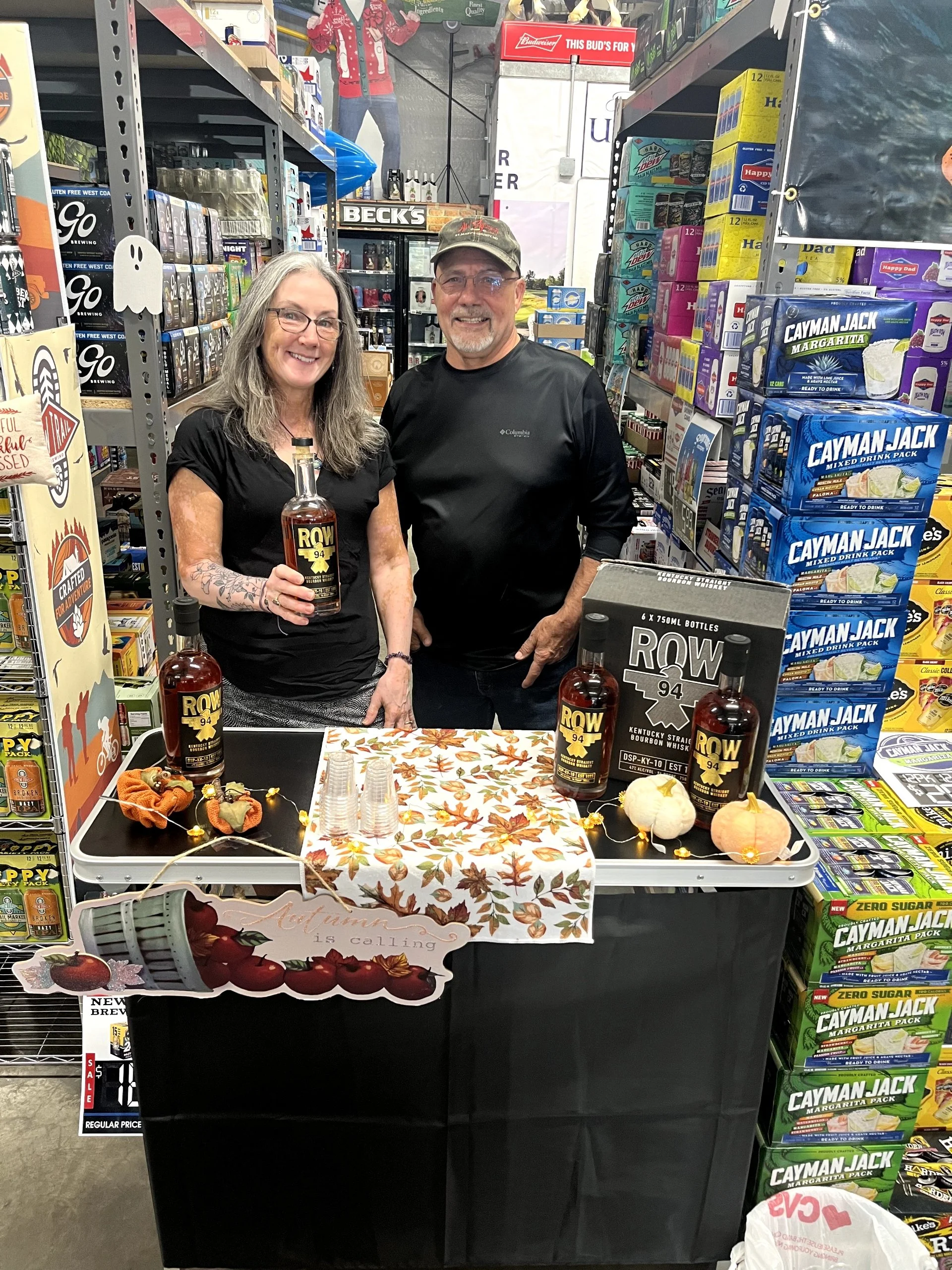 Two people stand behind a table displaying bottles of ROW94 bourbon whiskey, with a woman holding a bottle and a man smiling. The table is decorated with autumn leaves and small pumpkins, and is set up in a grocery store aisle with shelves stocked wi