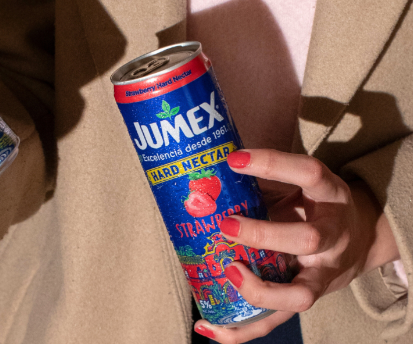 A person's hand with red painted nails holding a can of Jumex strawberry hard nectar drink. The can is blue with red and yellow accents and features an image of a strawberry.