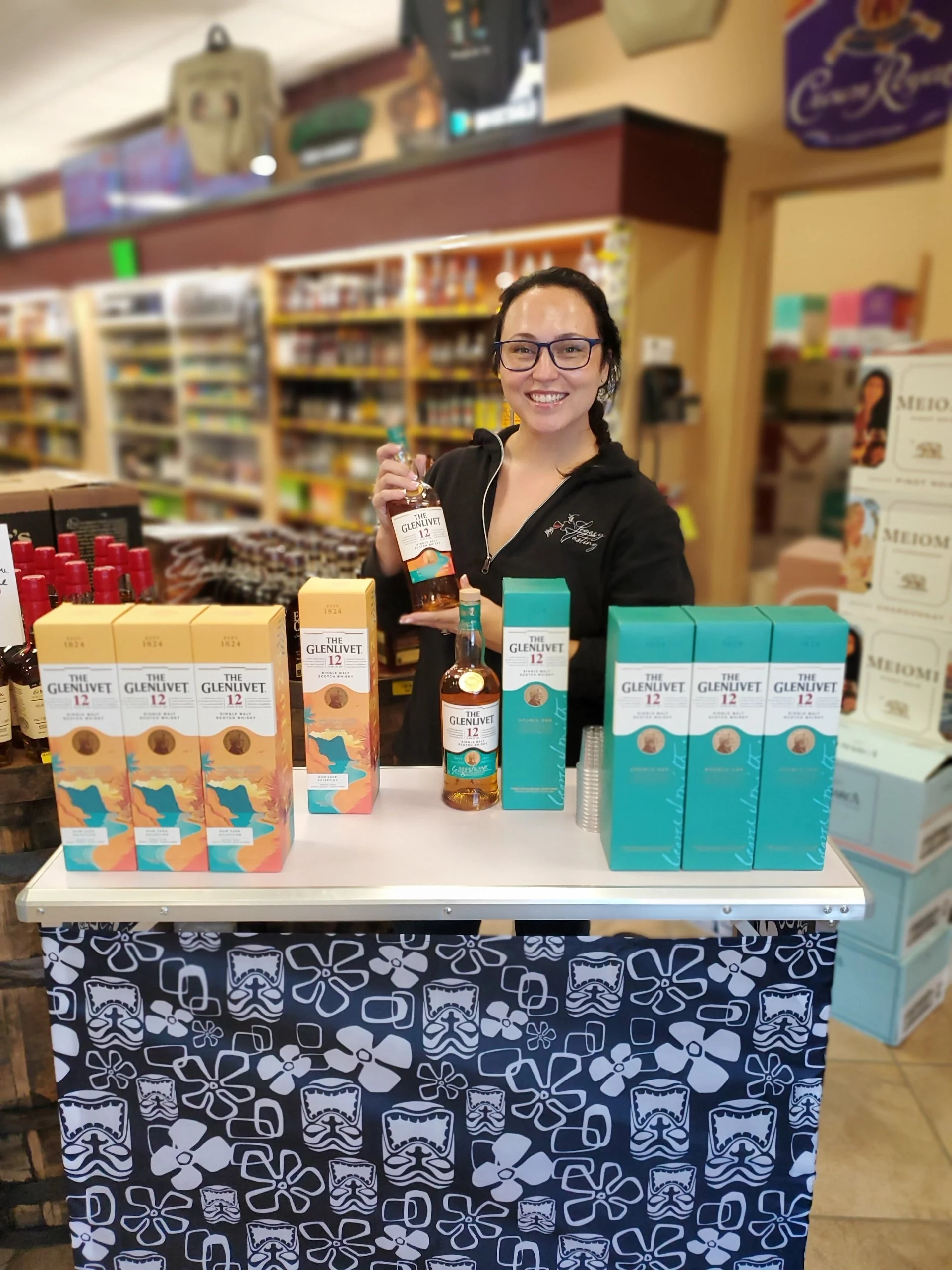 Woman smiling at a whisky tasting booth, holding a bottle of The Glenlivet 12-year-old single malt Scotch whisky. The booth has bottles and boxes of The Glenlivet whisky on display, with a backdrop featuring shelves of various products in a store or 
