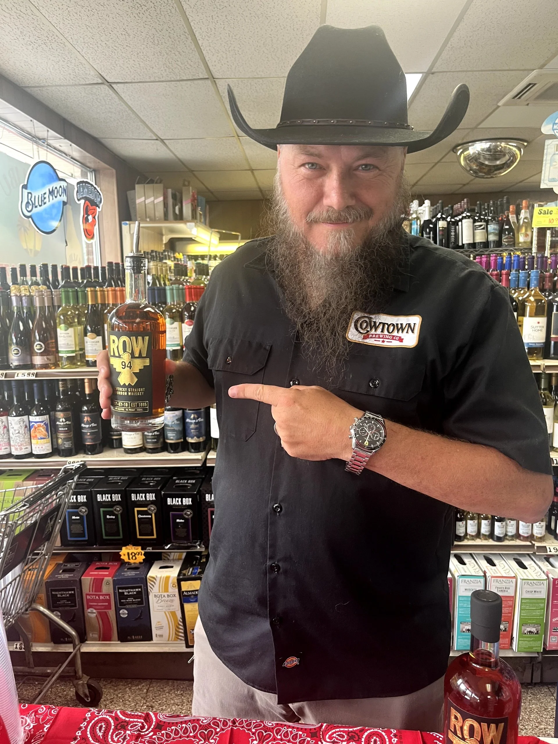 A man wearing a black cowboy hat, black shirt, and a wristwatch is holding a bottle of ROW whiskey, pointing at it with his finger. He is standing inside a liquor store with shelves of bottles behind him.