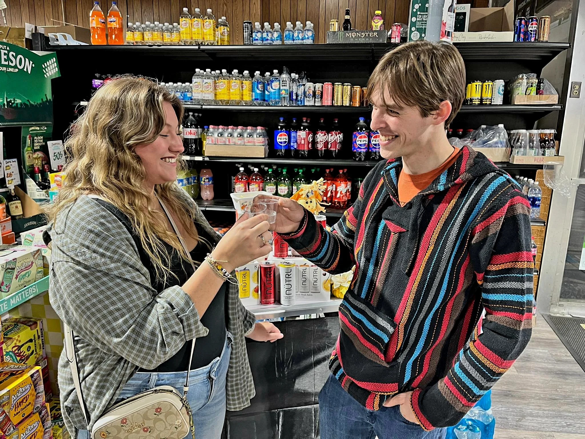 A young man and woman smiling and exchanging a shot glass in a convenience store aisle with drinks and snacks on shelves behind them.