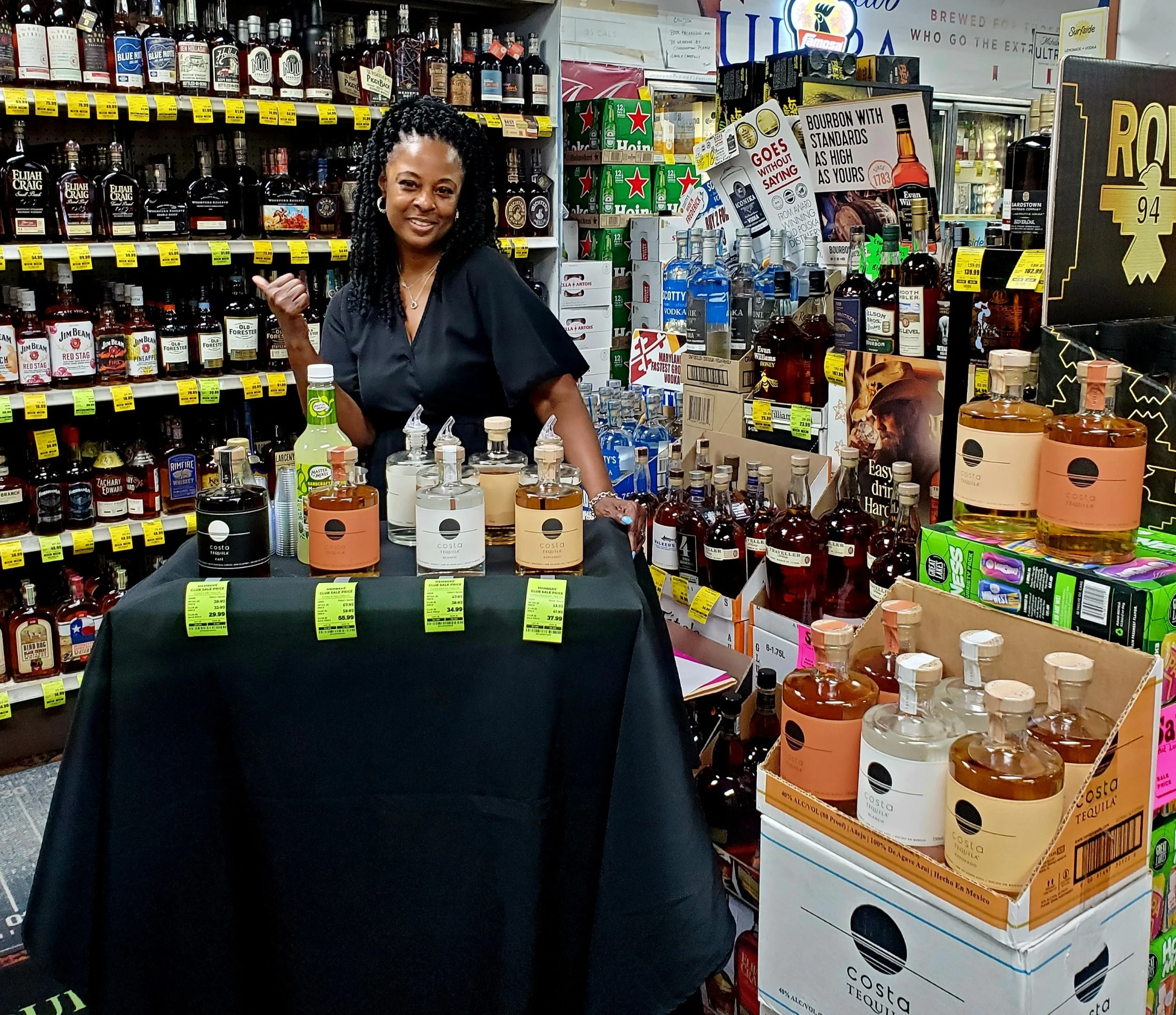 A woman standing behind a table with various bottles of tequila on display in a liquor store aisle.