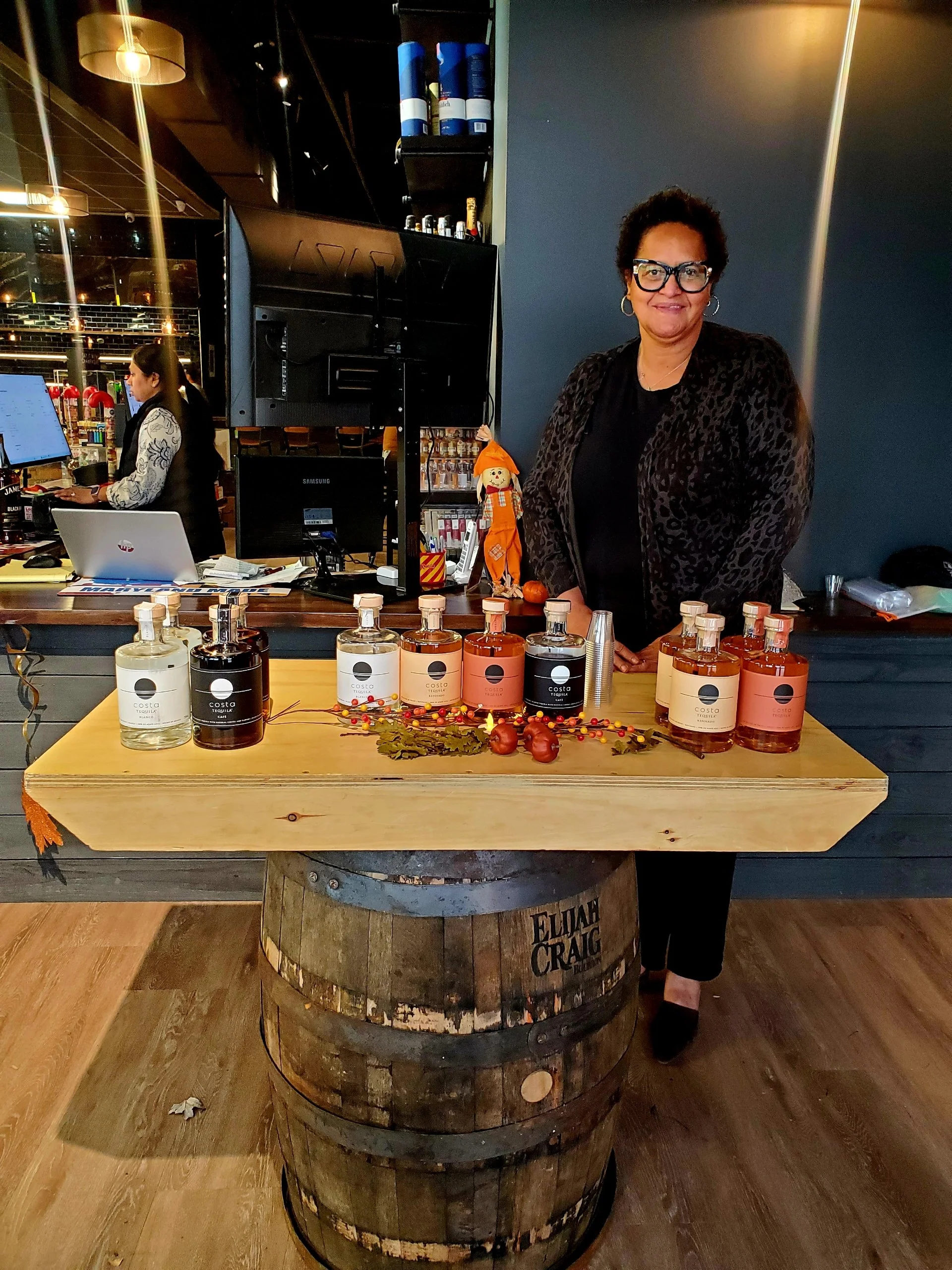 Woman standing behind a display table with bottles of tequila at a bar or restaurant, decorated with autumn-themed items, with a bartender and computer in the background.