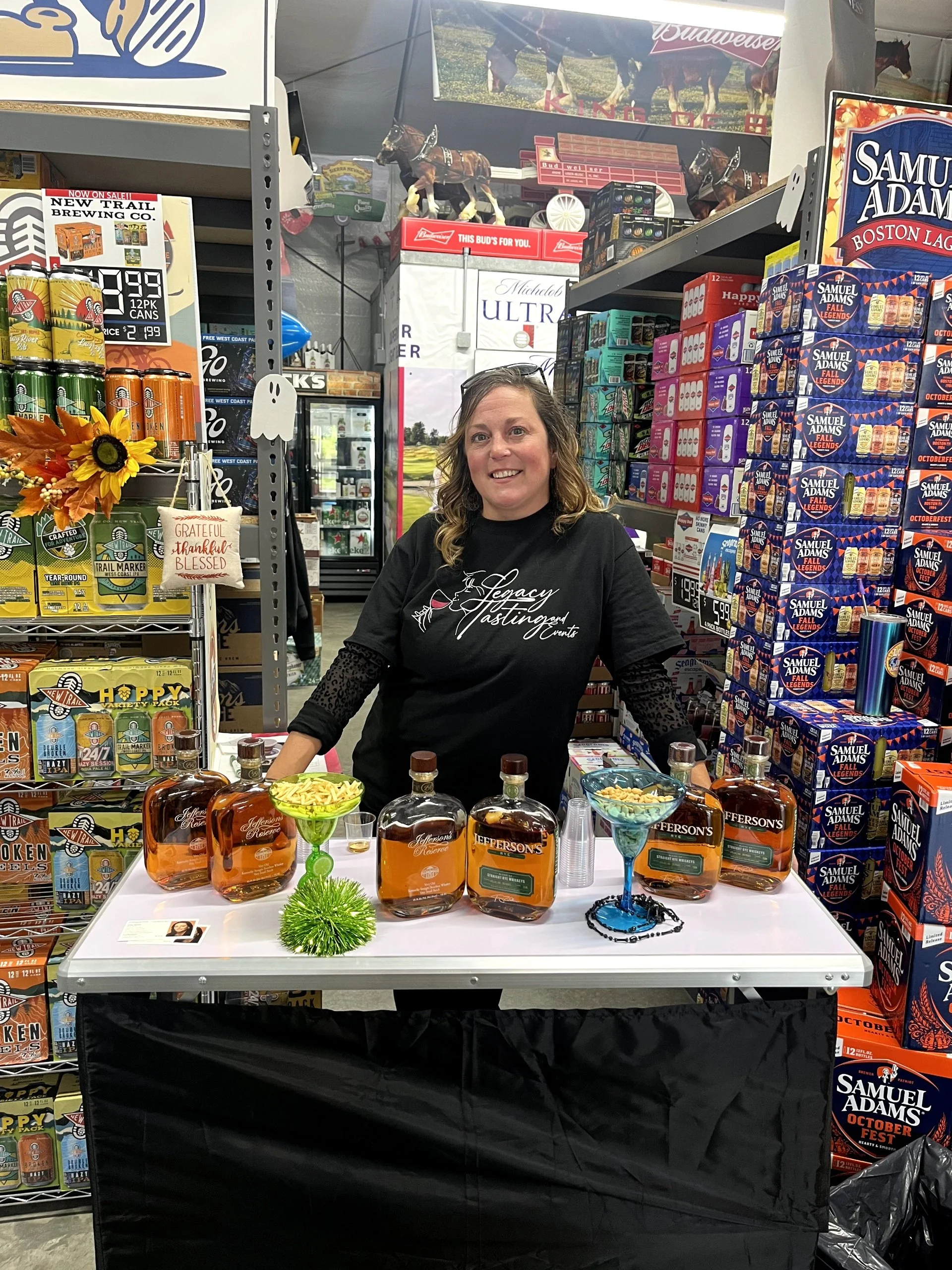 A woman standing behind a table displaying bottles of Jefferson's bourbon whiskey at a store. The table has bowls with snacks and decorative items. The store shelves behind are stocked with snack and beverage boxes, and a poster of horses in a field 