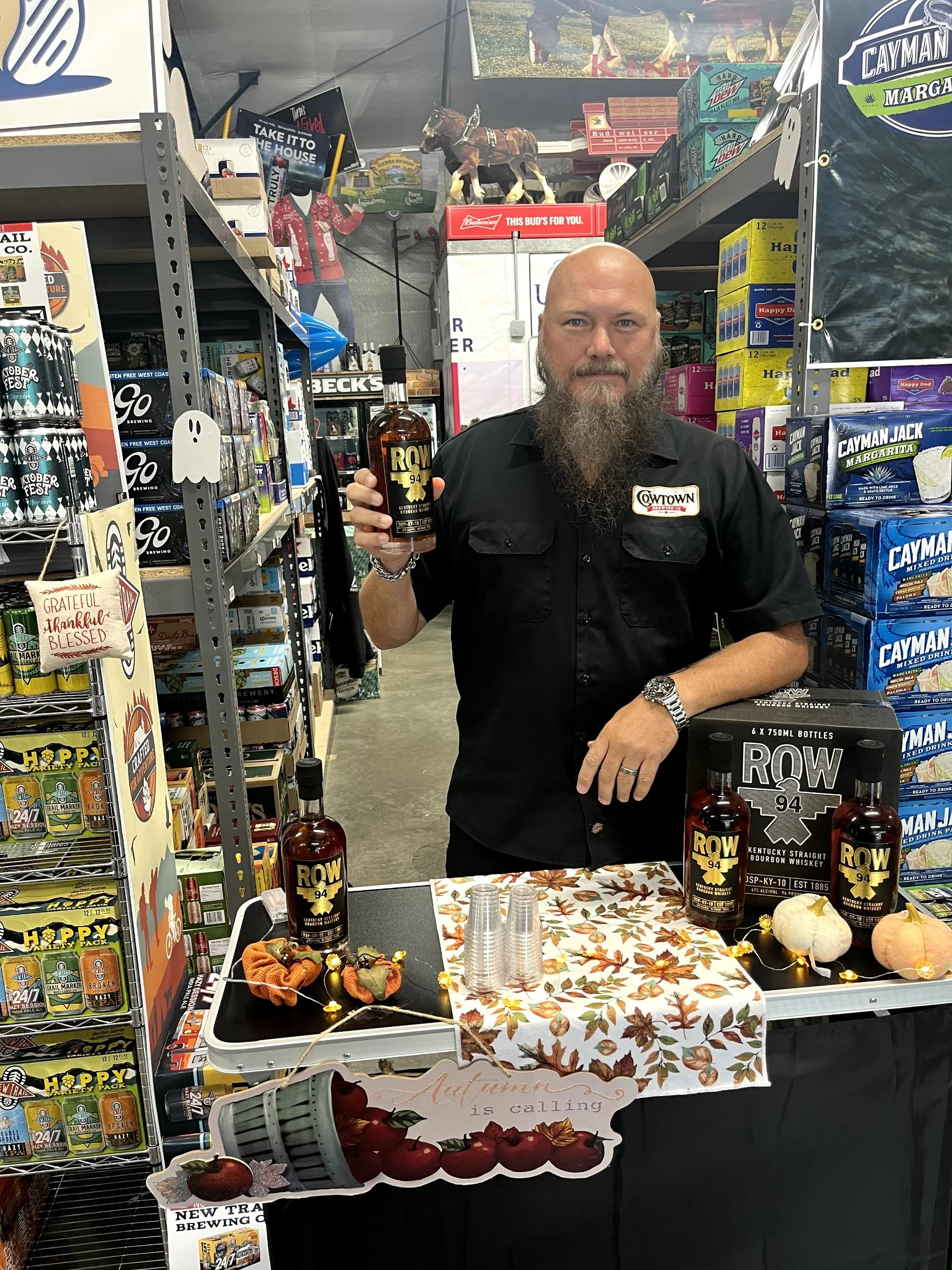 A man with a beard and a shaved head standing behind a display table with bottles of bourbon whiskey, holding a bottle in his right hand. The table has decorative autumn pumpkins, a tablecloth with fall leaves, and plastic cups. The man is wearing a 