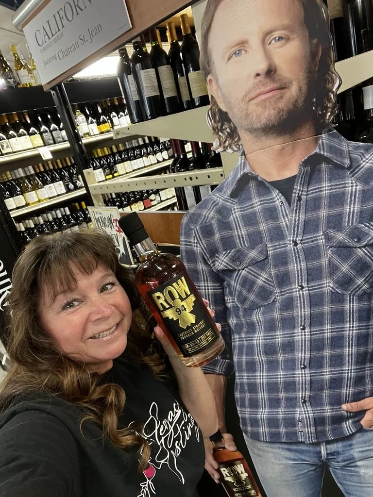Woman holding a bottle of ROW 94 whiskey next to a standee of a man at a wine store.