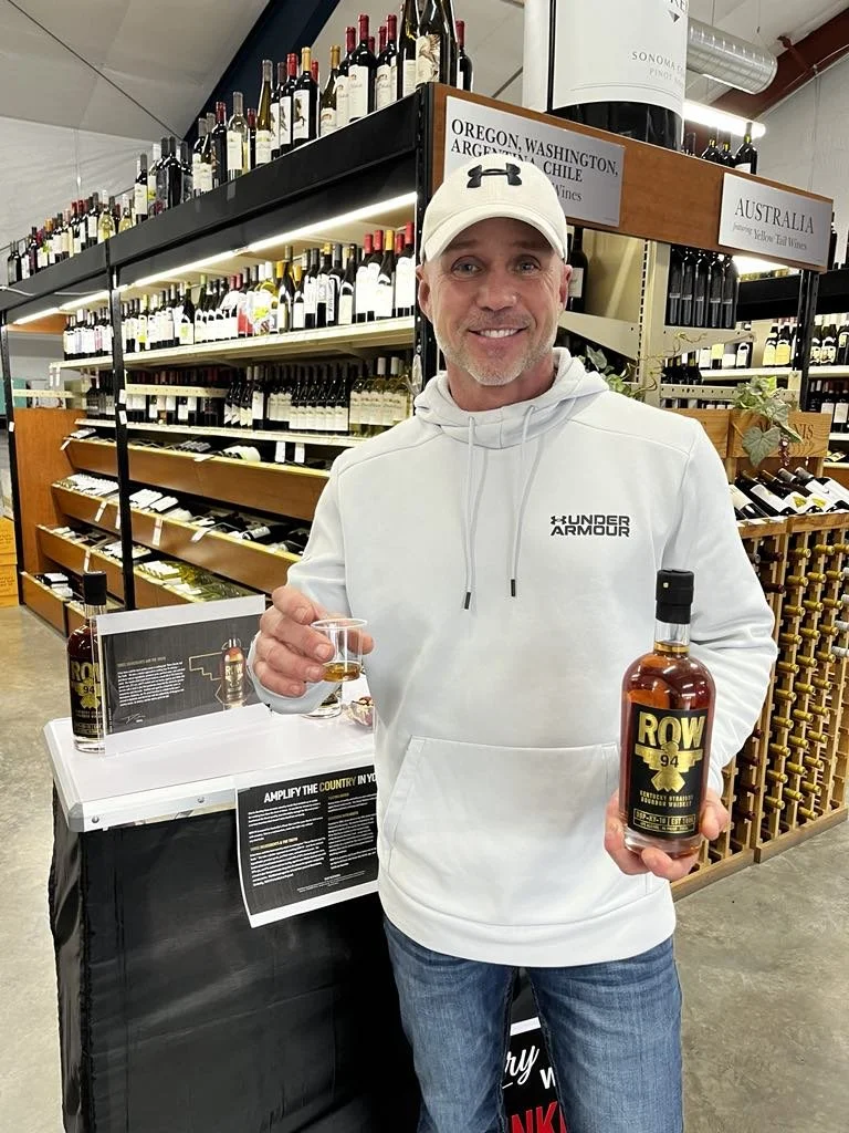 A man smiling and holding a glass of whiskey in one hand and a bottle labeled ROW 94 in the other, standing in a wine store with shelves of wine bottles behind him.