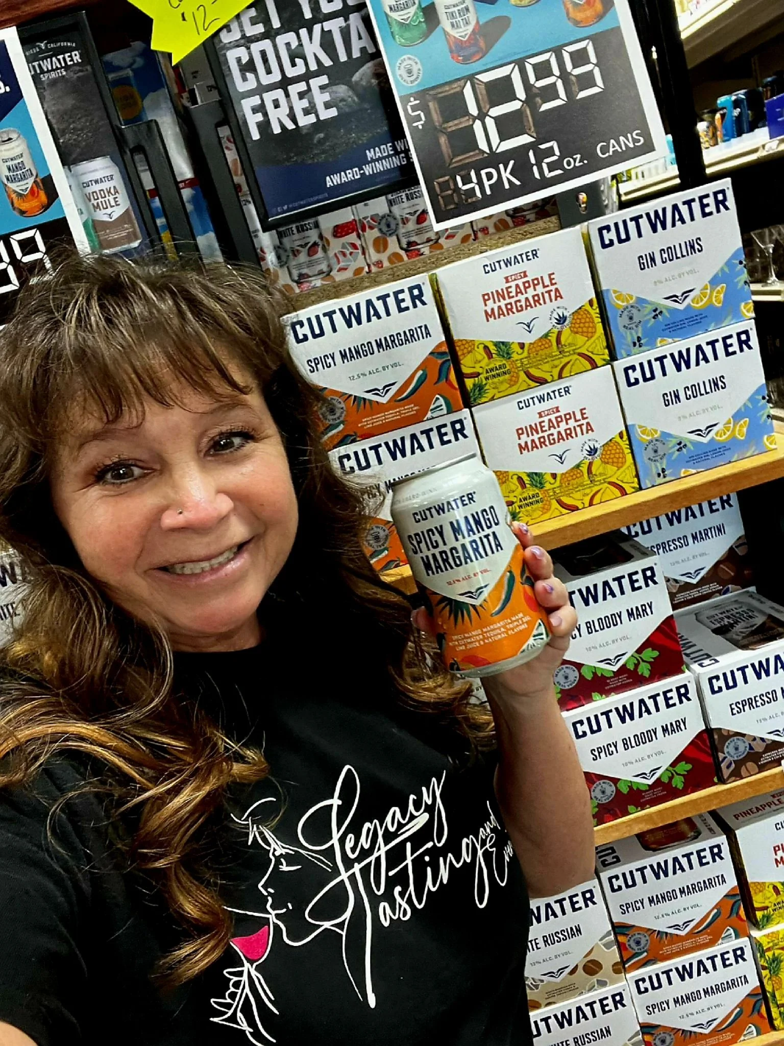 Woman with wavy brown hair smiling and holding a can of Cutwater Spicy Mango Margarita in a store aisle with shelves of canned drinks and boxes of Cutwater beverages behind her.