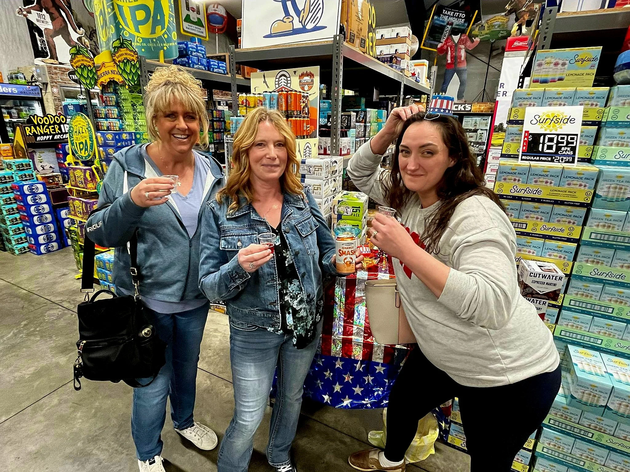 Three women posing inside a store, with one of them holding a shot glass, facing the camera. The woman on the right is adjusting a hat on her head. They are near a display of juice boxes and snacks, with store shelves and signs visible in the backgro