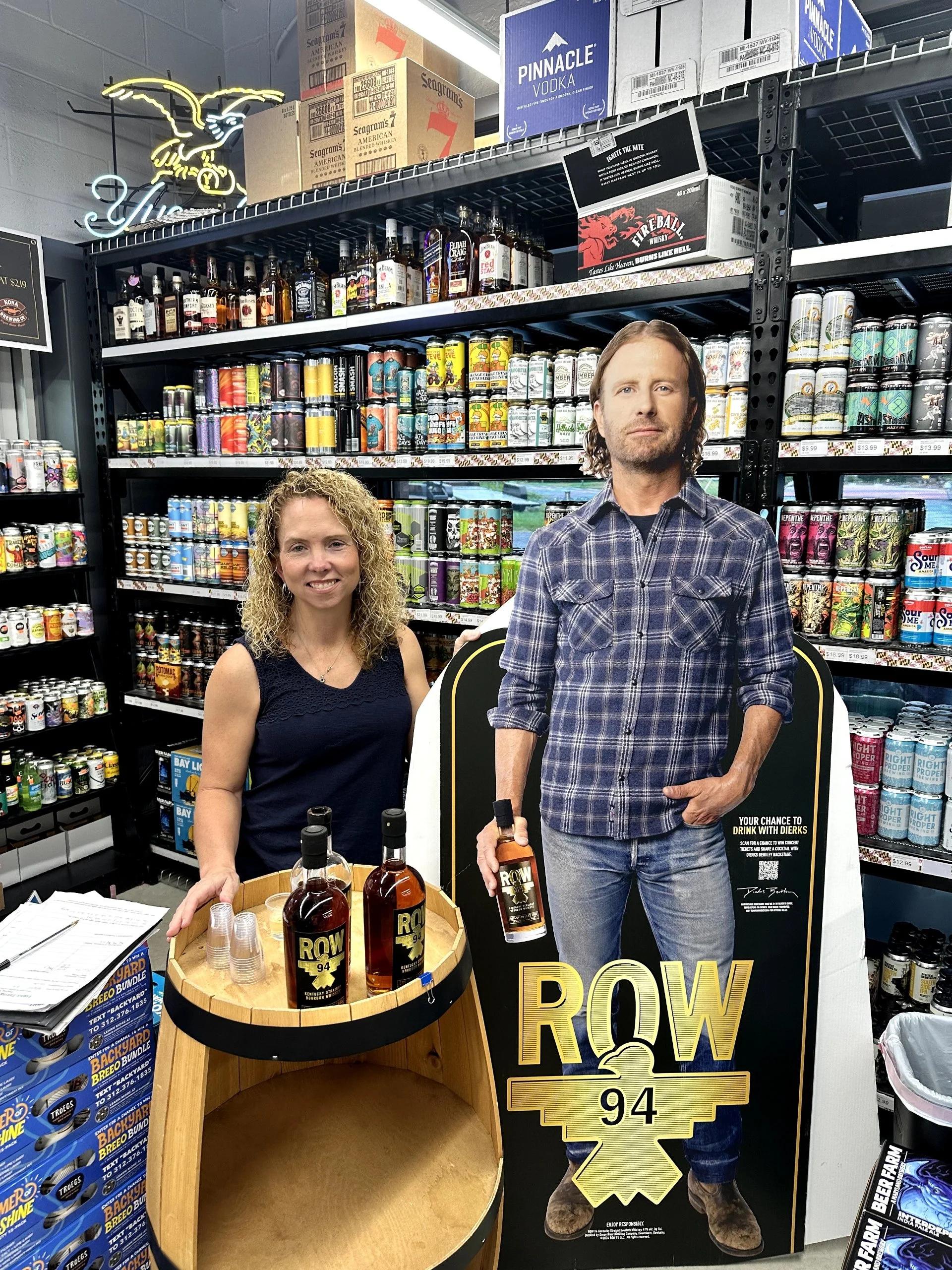 A woman standing behind a display table with bottles of Row 94 whiskey and plastic shot glasses, in a store aisle filled with craft beers and spirits on shelves. A life-size cardboard cutout of a man holding a bottle of Row 94 whiskey is next to her.