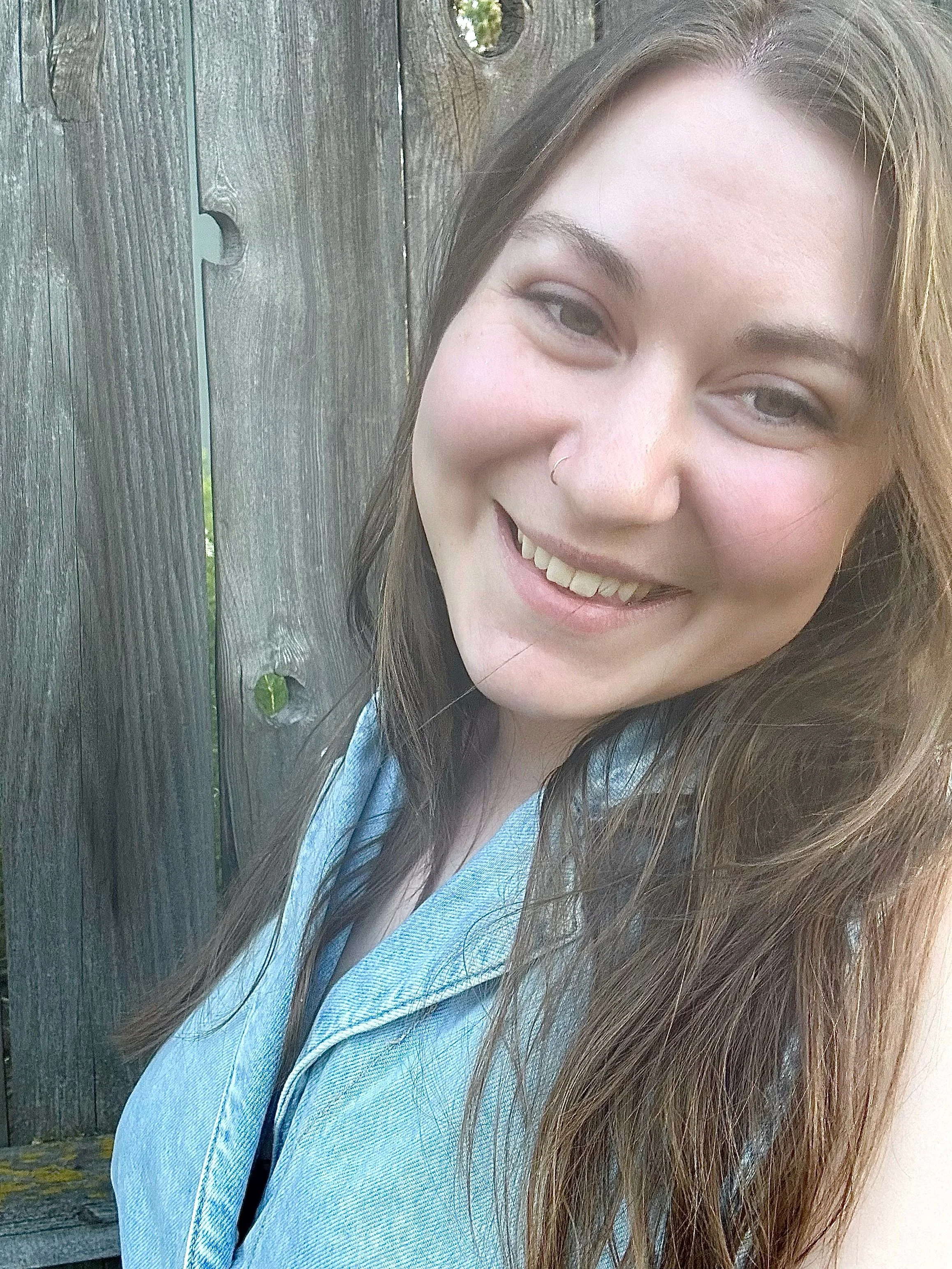 A young woman with long brown hair and a nose piercing smiling outdoors in front of a wooden fence.