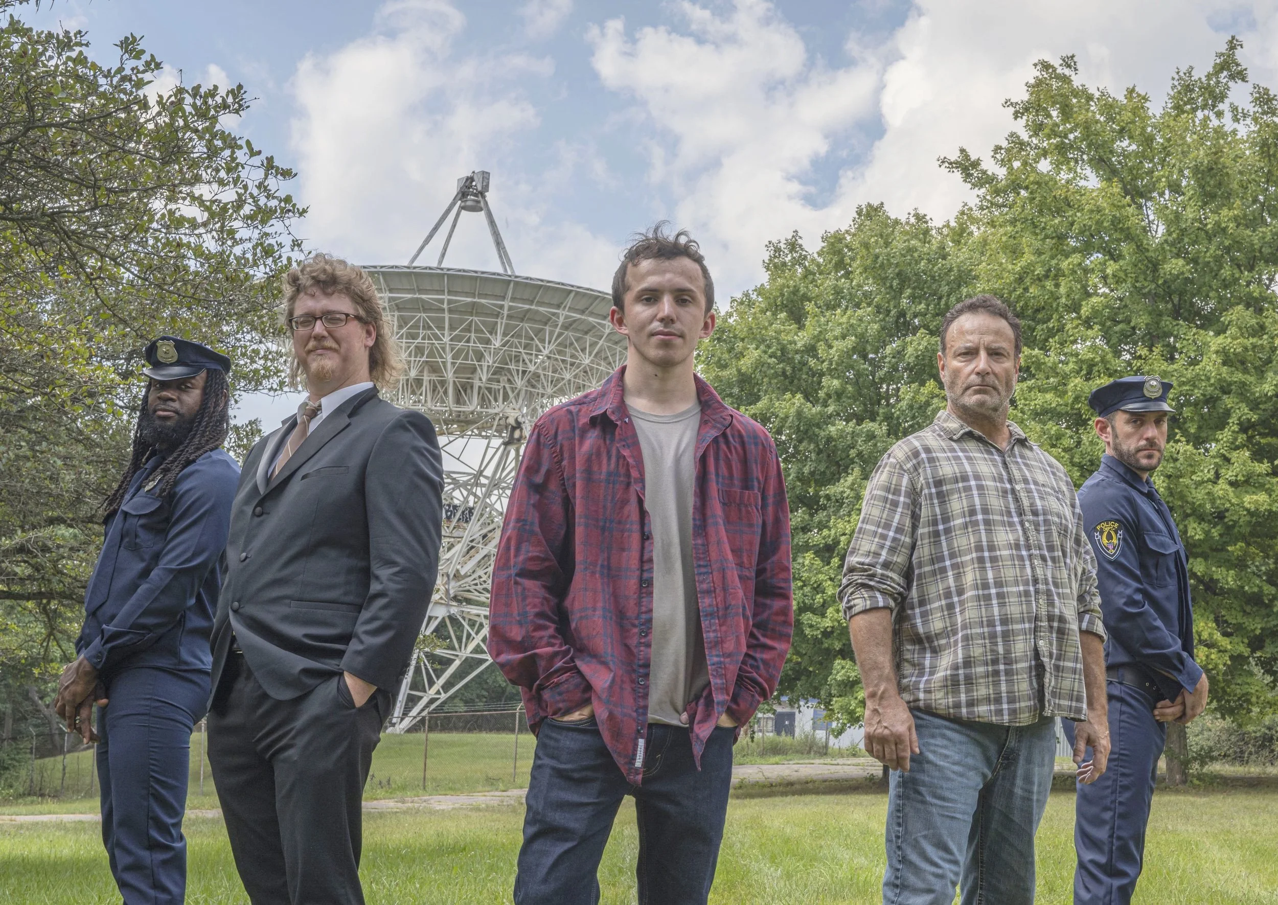 Five people standing outdoors in front of a large satellite dish, with trees and clouds in the background. The group includes two police officers, a man in a suit, a young man in casual clothing, and an older man in a plaid shirt.