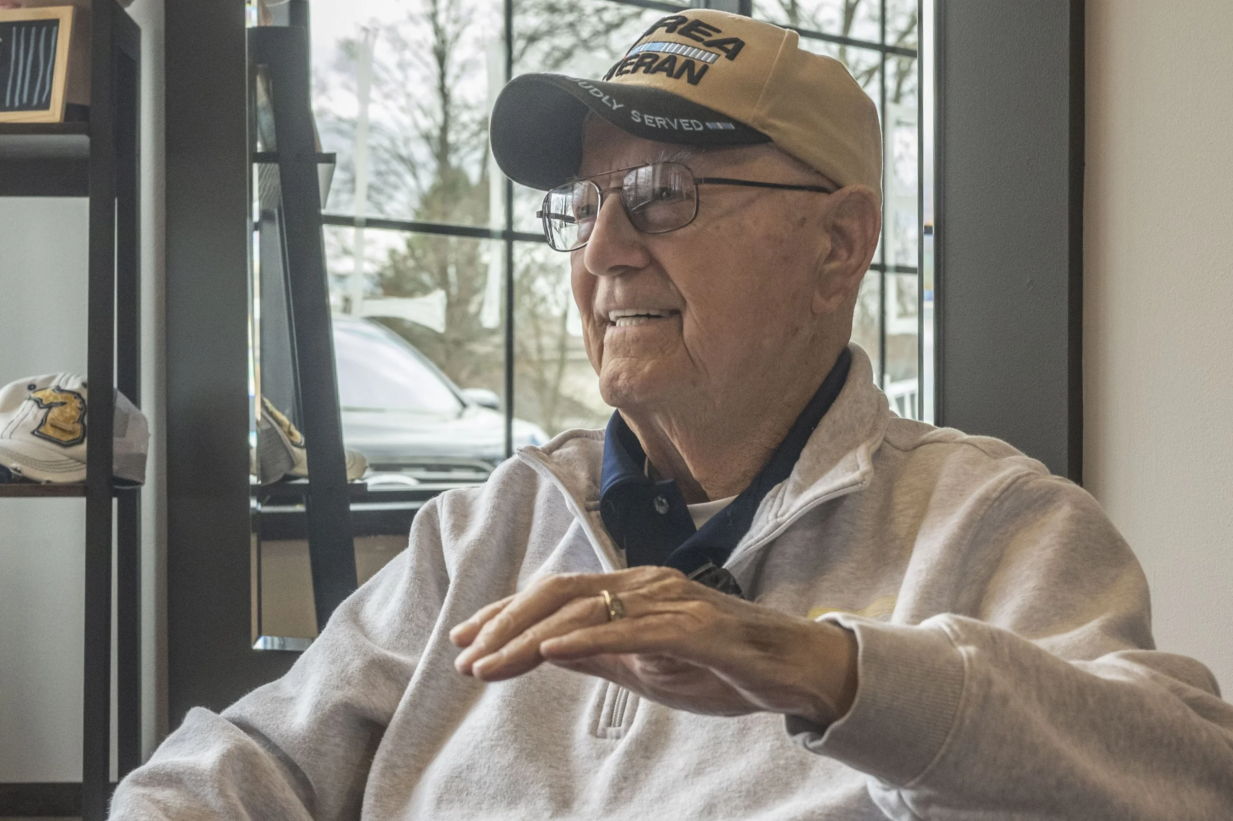An elderly man wearing glasses, a beige cap, and a light gray zip-up jacket sitting indoors, smiling and gesturing with his hand. Behind him a large window shows trees and parked cars outside.