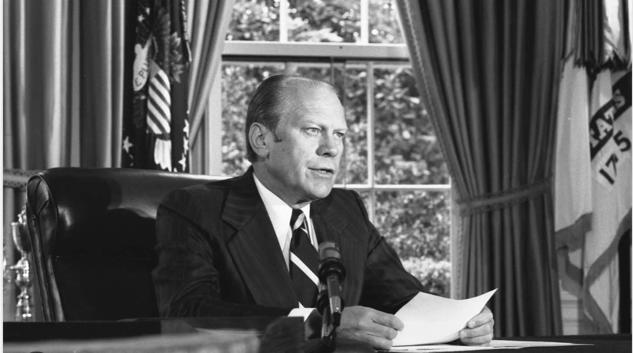 A man in a suit sitting at a desk with a microphone, holding papers, in a room with windows and curtains, with U.S. flags in the background.