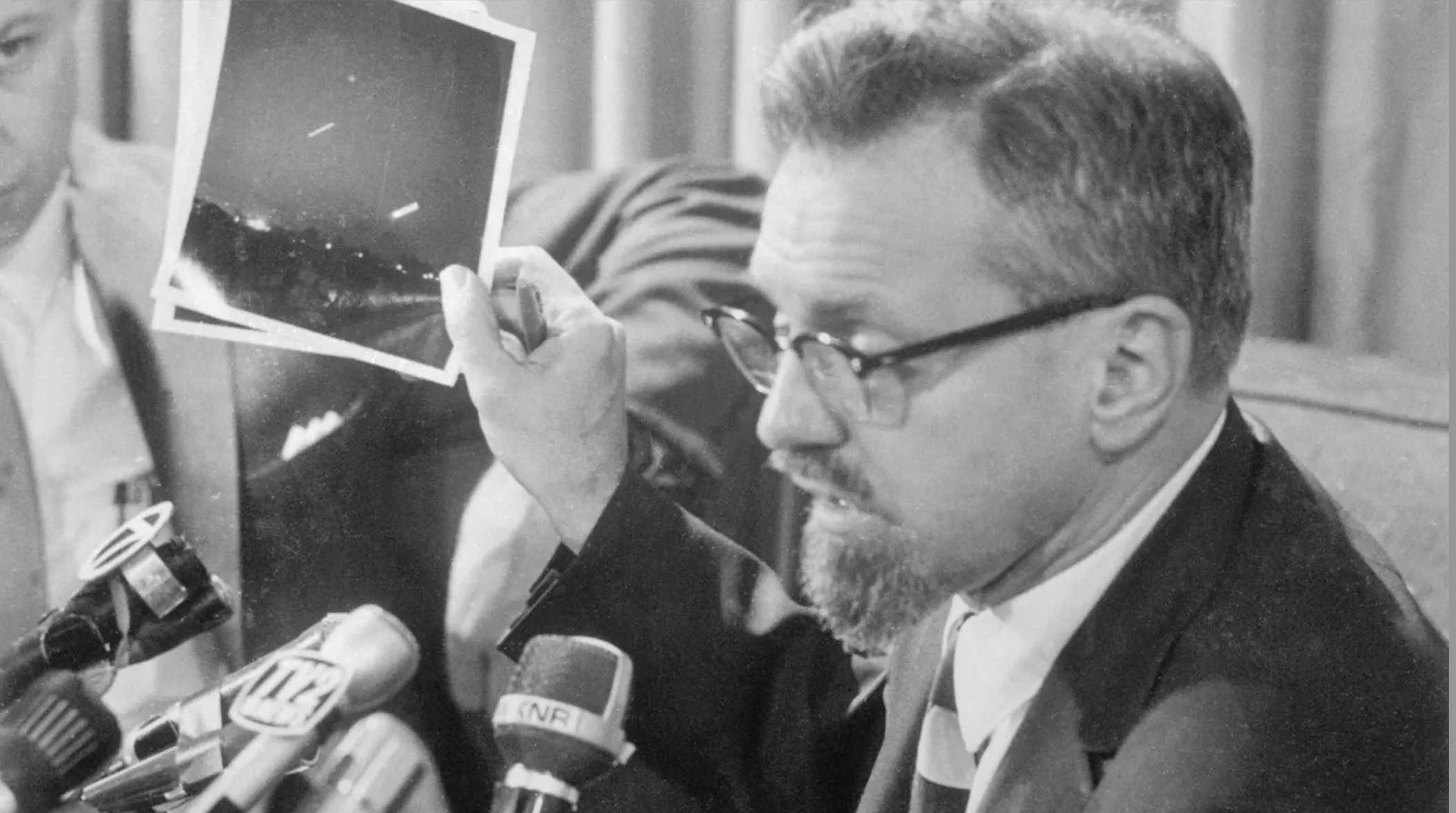 A black and white photograph of a man with glasses, a mustache, and a beard, sitting at a table with microphones in front of him. He is looking at a photograph held in his hand, which appears to depict a landscape at night with a snowy scene. The man is dressed in a suit and tie, and appears to be speaking or giving a statement.