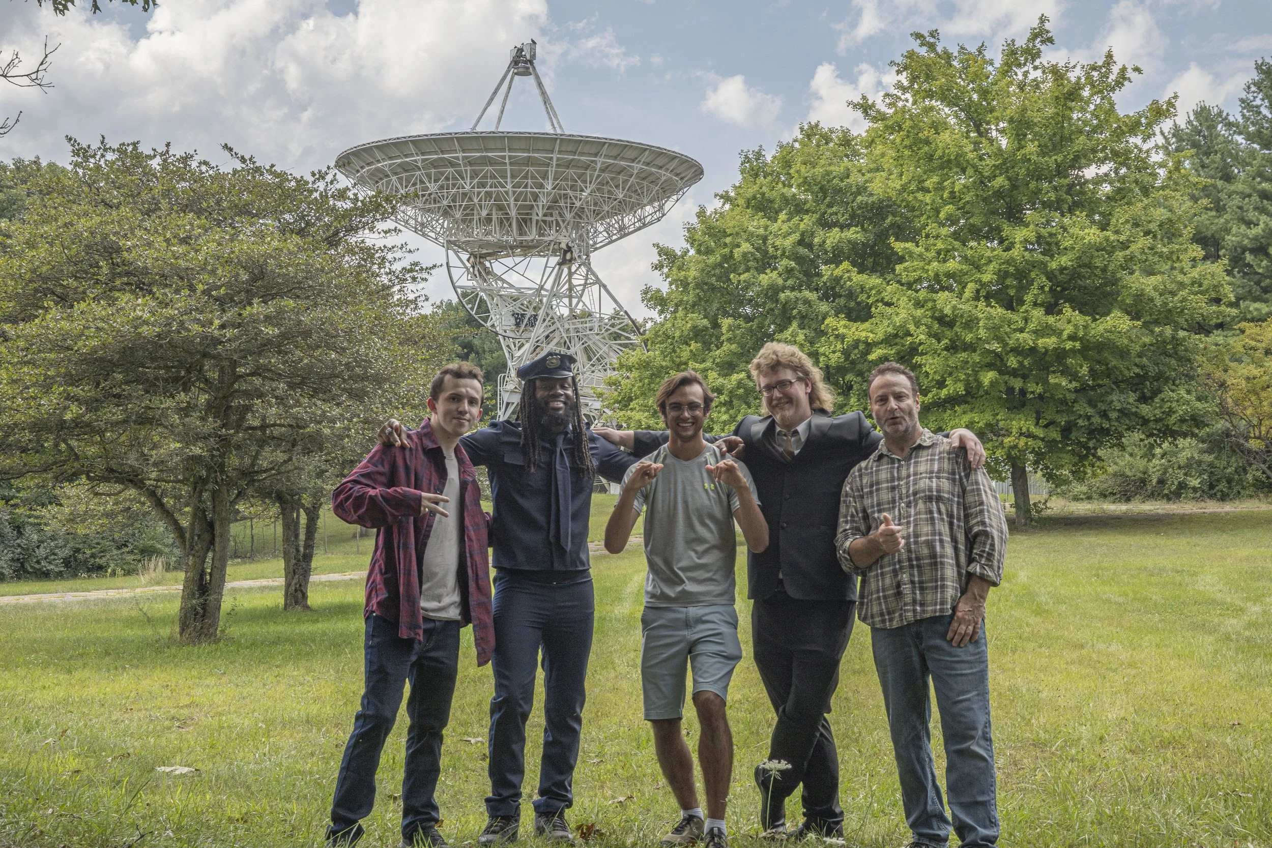 A group of five diverse people standing together outdoors in a grassy park with trees and a large radio telescope in the background.