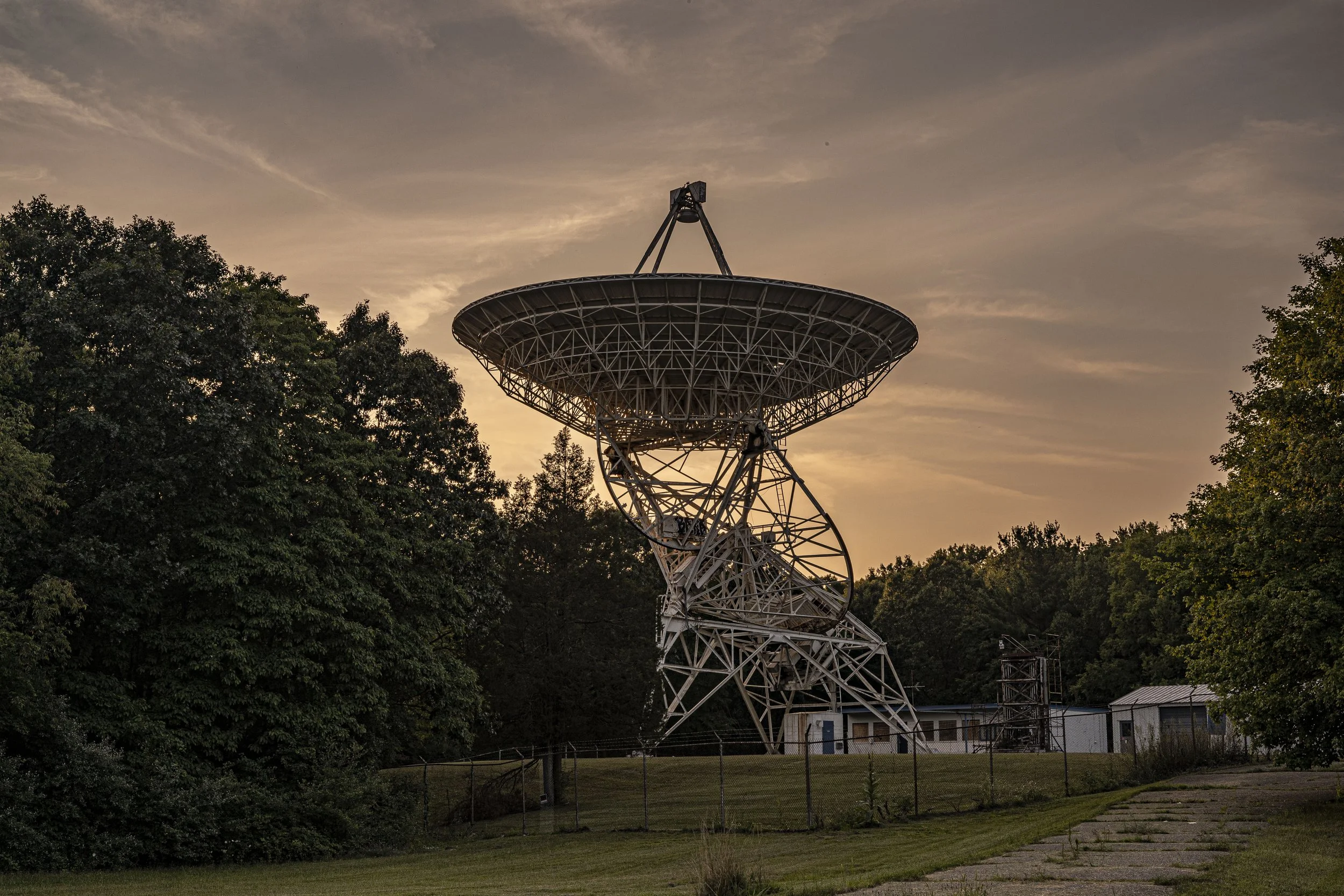 A large radio telescope dish on a metal structure surrounded by trees at sunset.