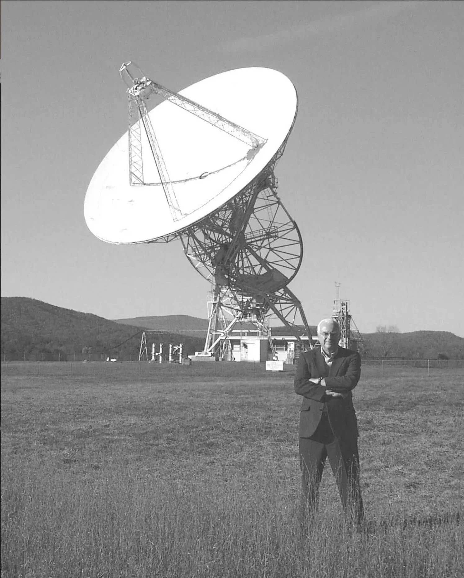 A man in a suit standing with arms crossed in front of a large satellite dish on a grassy field, with hills in the background.